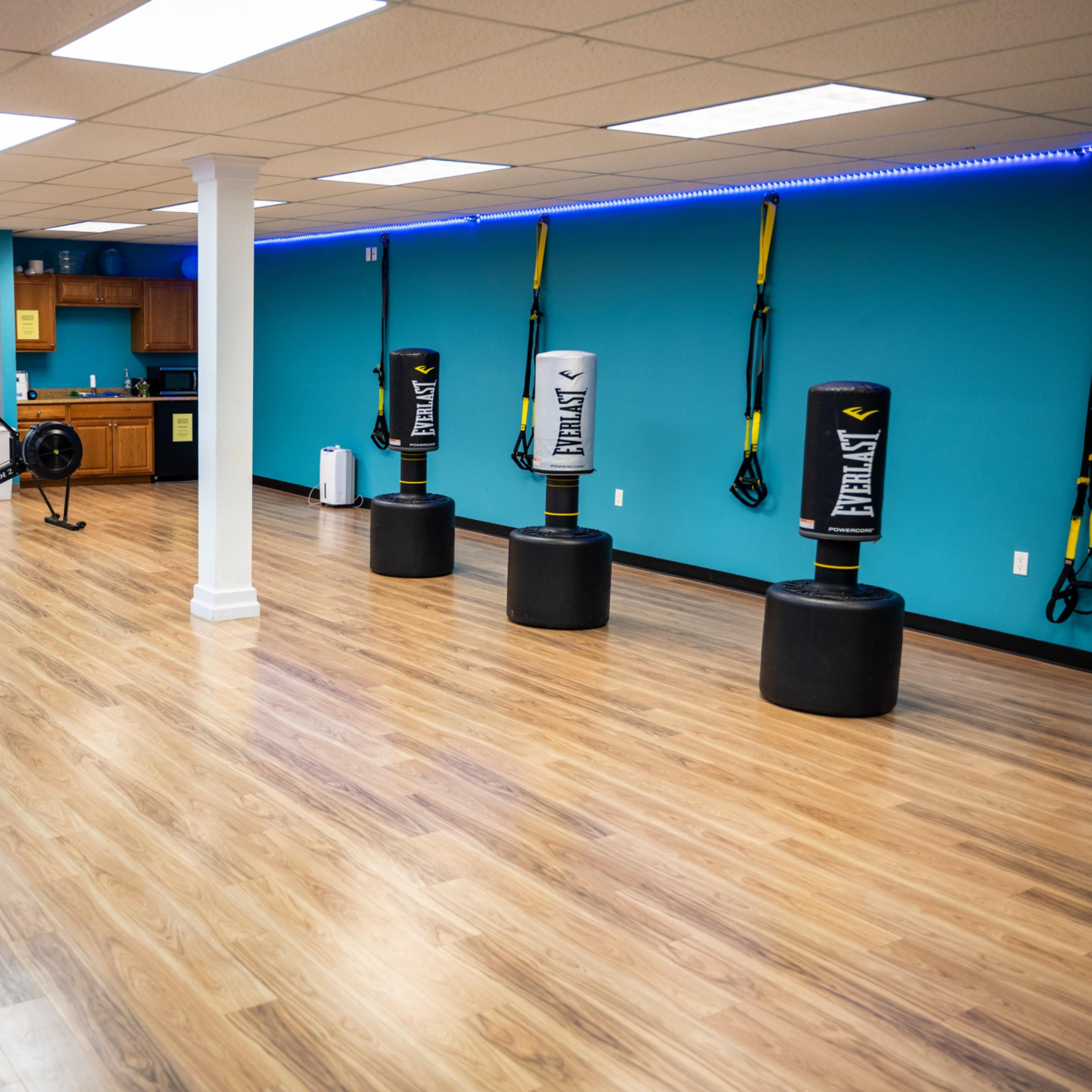 A spacious fitness studio with wooden floors, blue walls, and various equipment including punching bags, exercise balls, and a kitchen area.