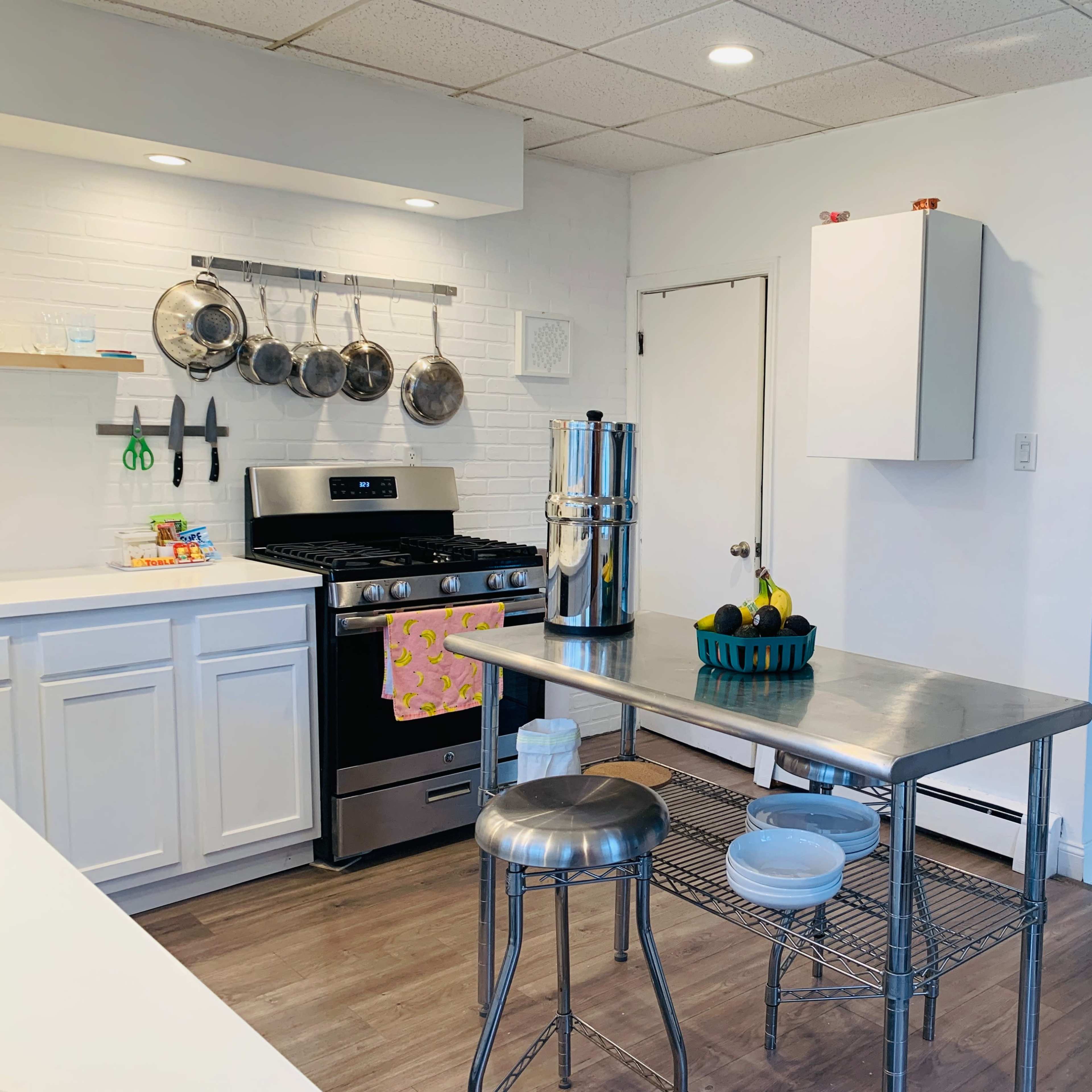 A modern kitchen features stainless steel appliances, a central metal table with a bowl of fruit, and white cabinetry against wooden flooring.