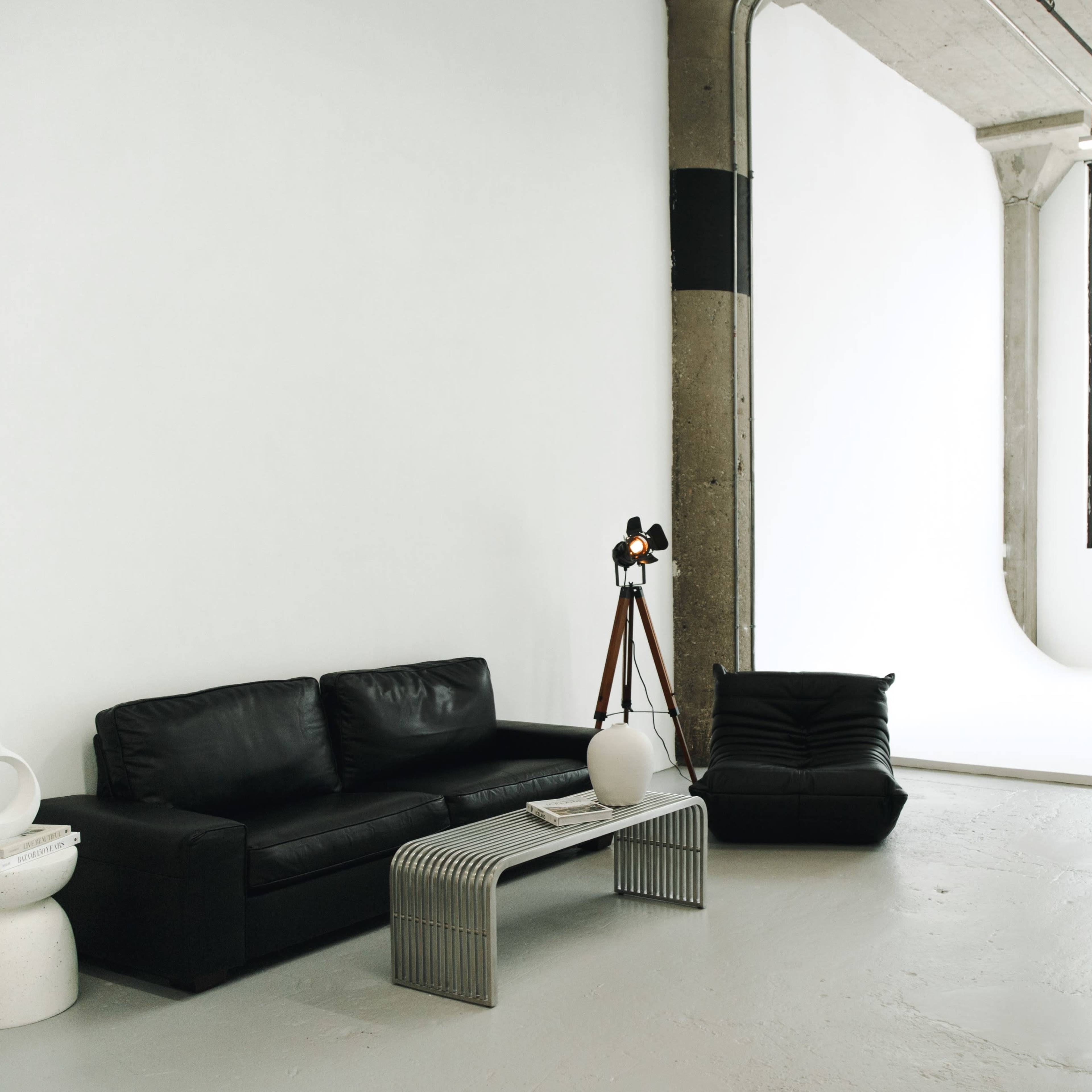 A spacious, well-lit interior featuring a black leather sofa, a modern table with a white vase, and large windows alongside a minimalist backdrop.