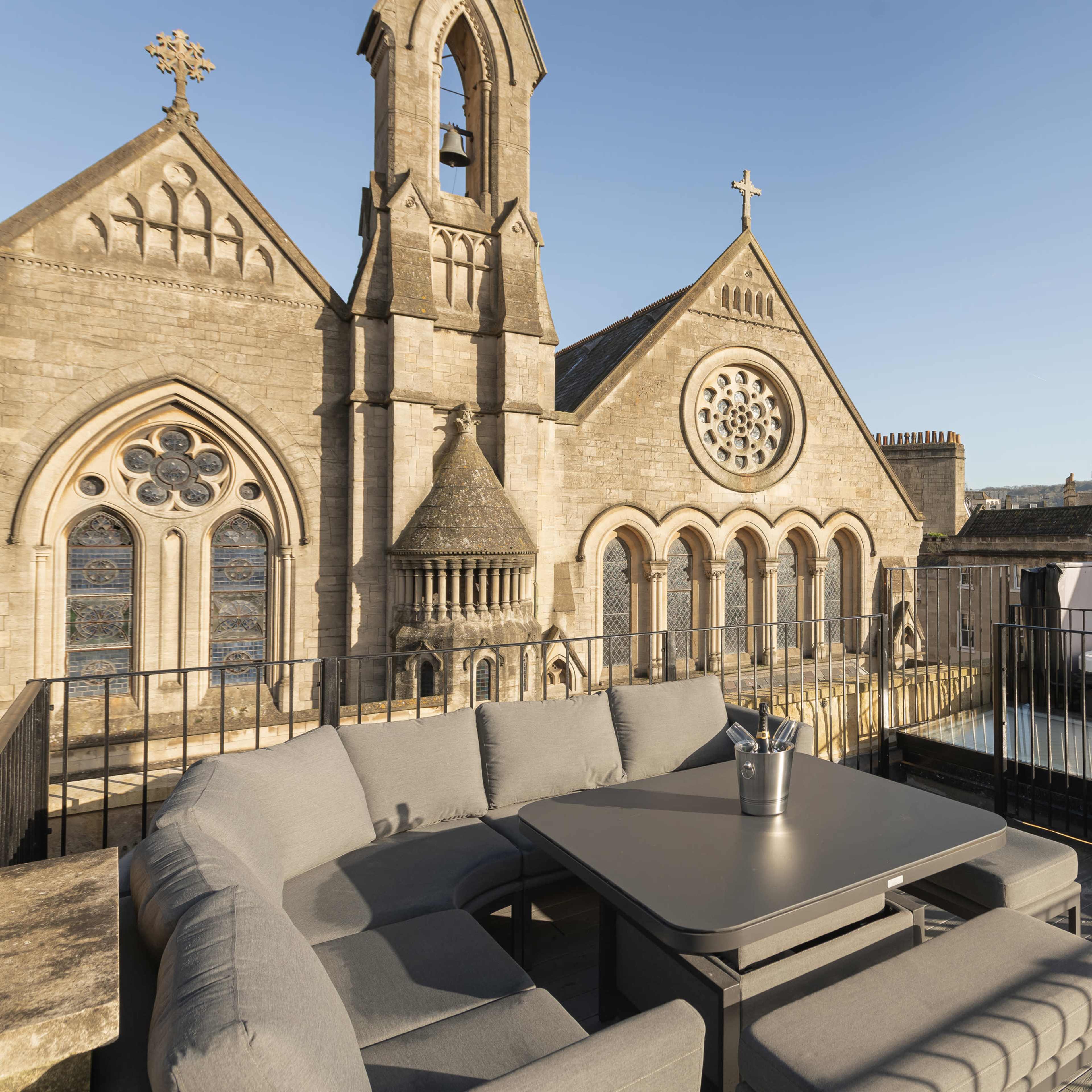 A rooftop terrace features a gray sectional sofa and table overlooking historic stone buildings with decorative architectural details.