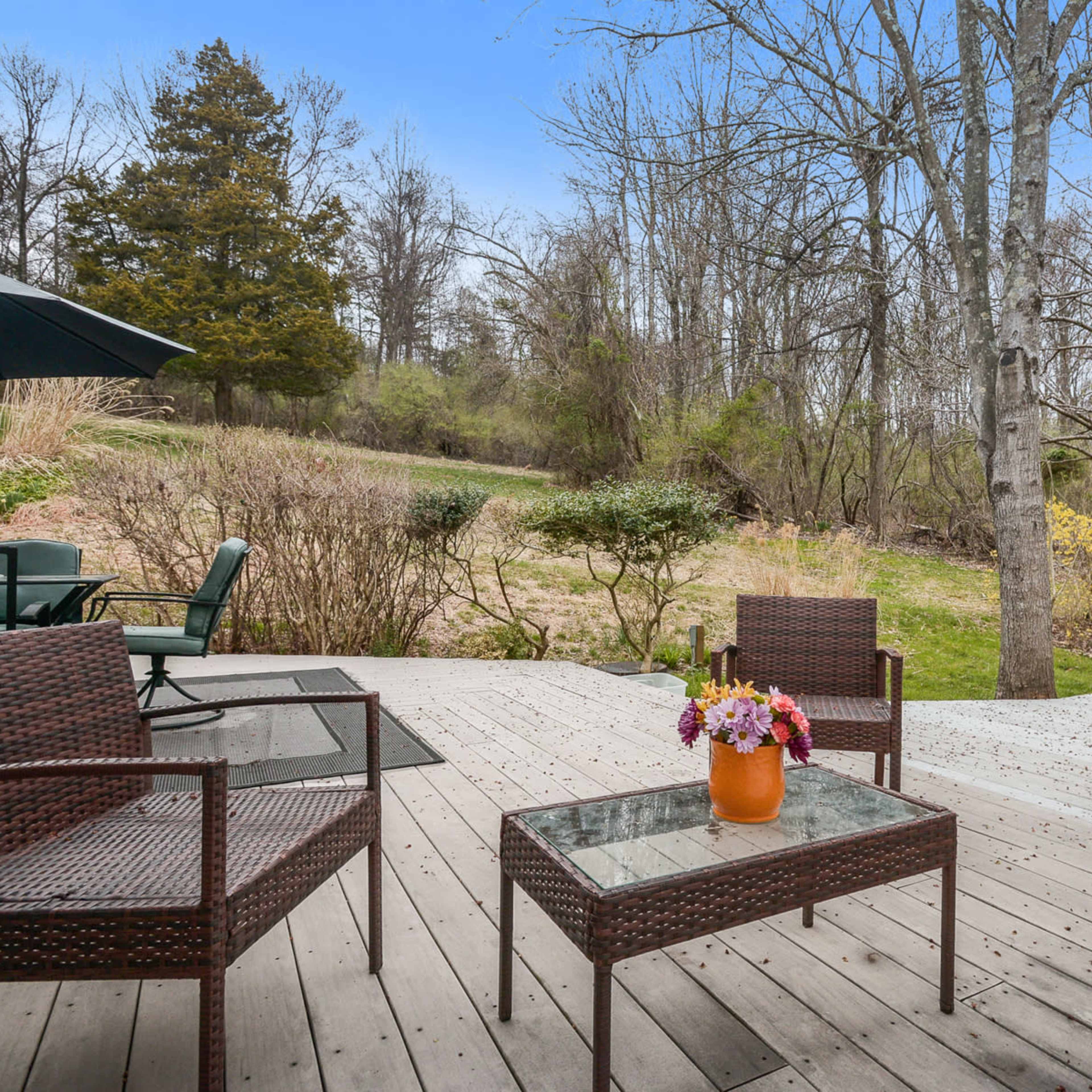 A wooden patio with two wicker chairs and a table holding a flower pot, overlooking a grassy yard with sparse trees.