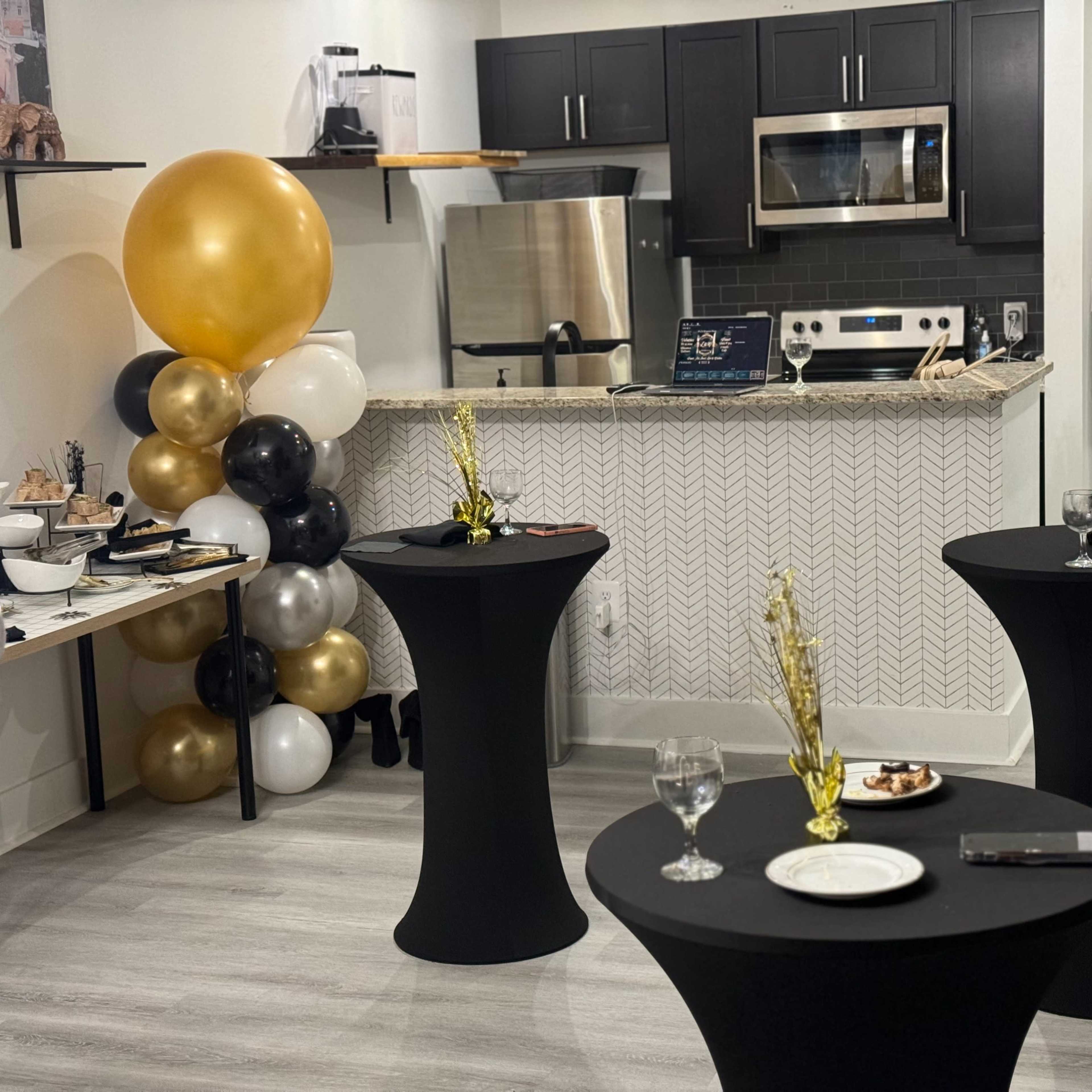 A modern kitchen with a small gathering area, featuring two black cocktail tables with plates and glasses, and festive balloons arranged in gold, black, and white nearby.