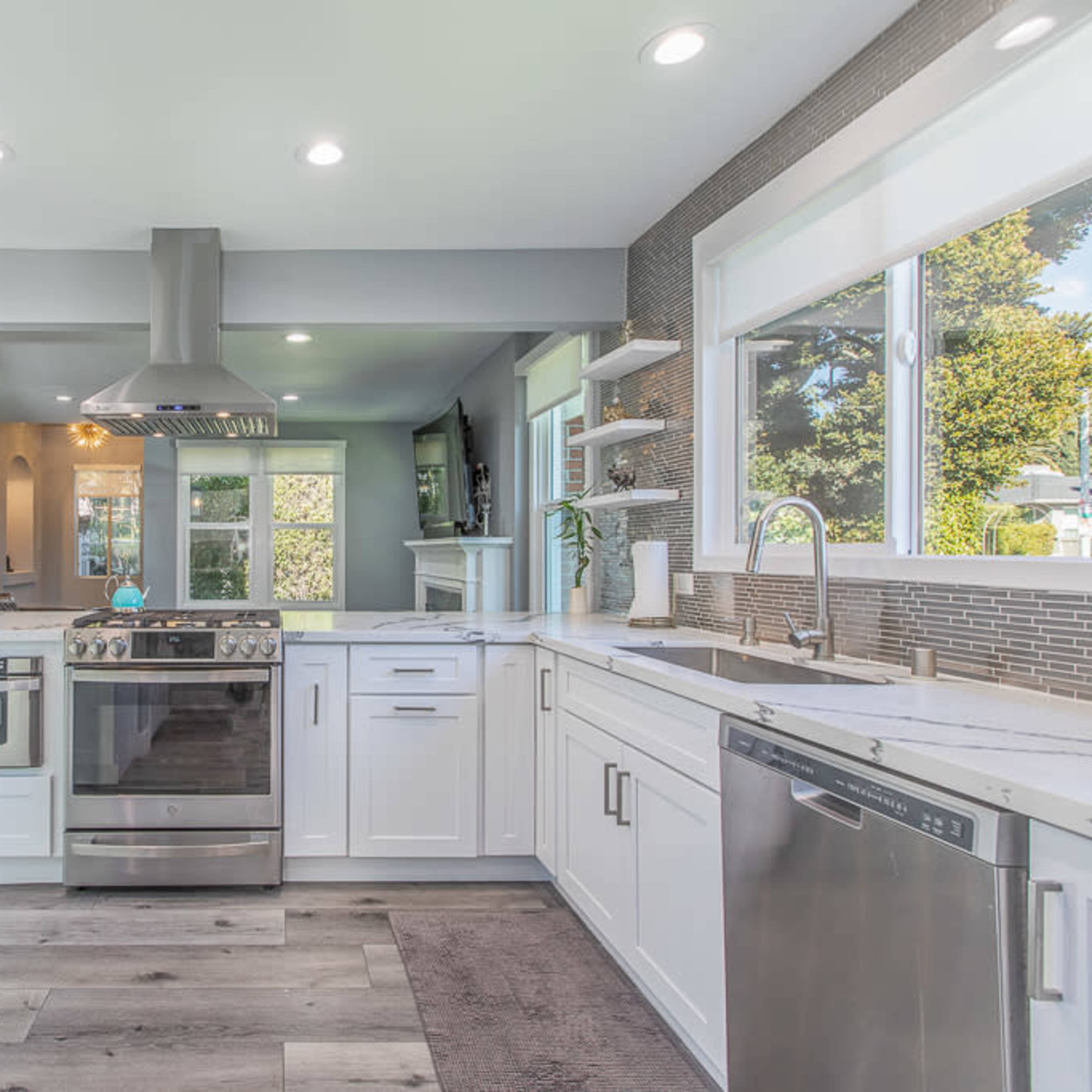 The kitchen features modern white cabinetry, stainless steel appliances, and a large window allowing natural light to flood the space.