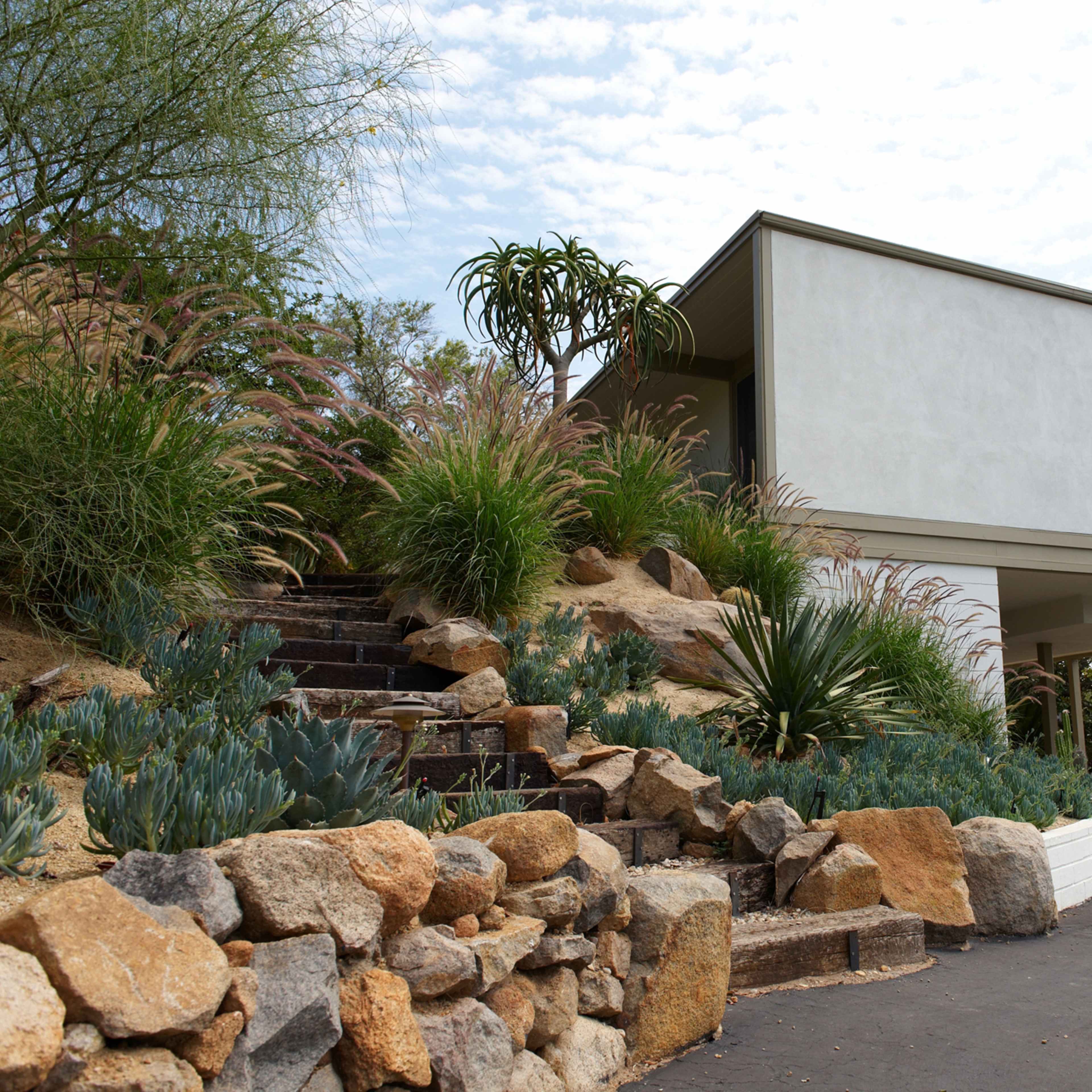 A modern two-story house is set on a landscaped hill with stone steps and various plants leading up to its entrance.