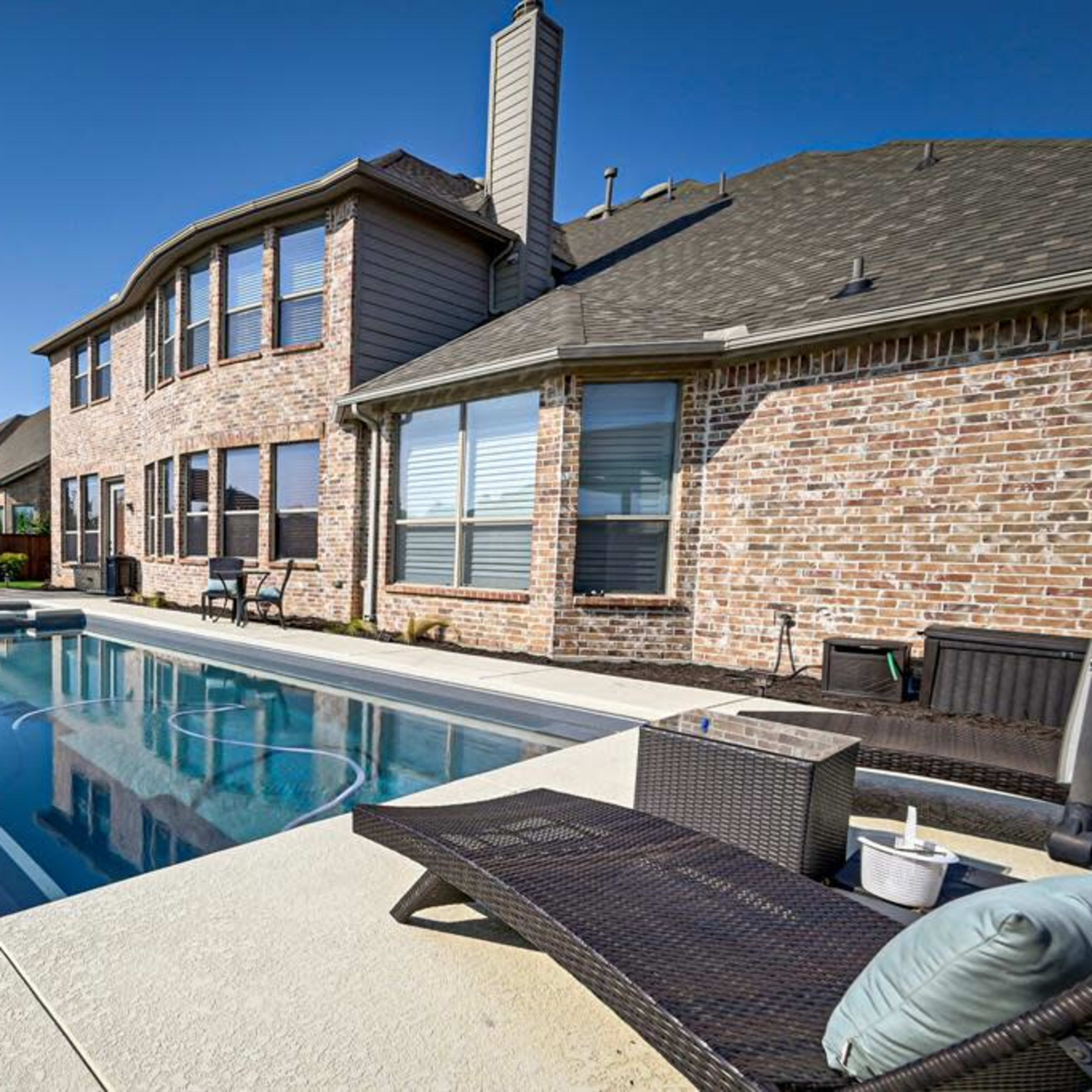 A backyard with a rectangular pool adjacent to a brick house, featuring a shaded seating area and landscaped greenery.