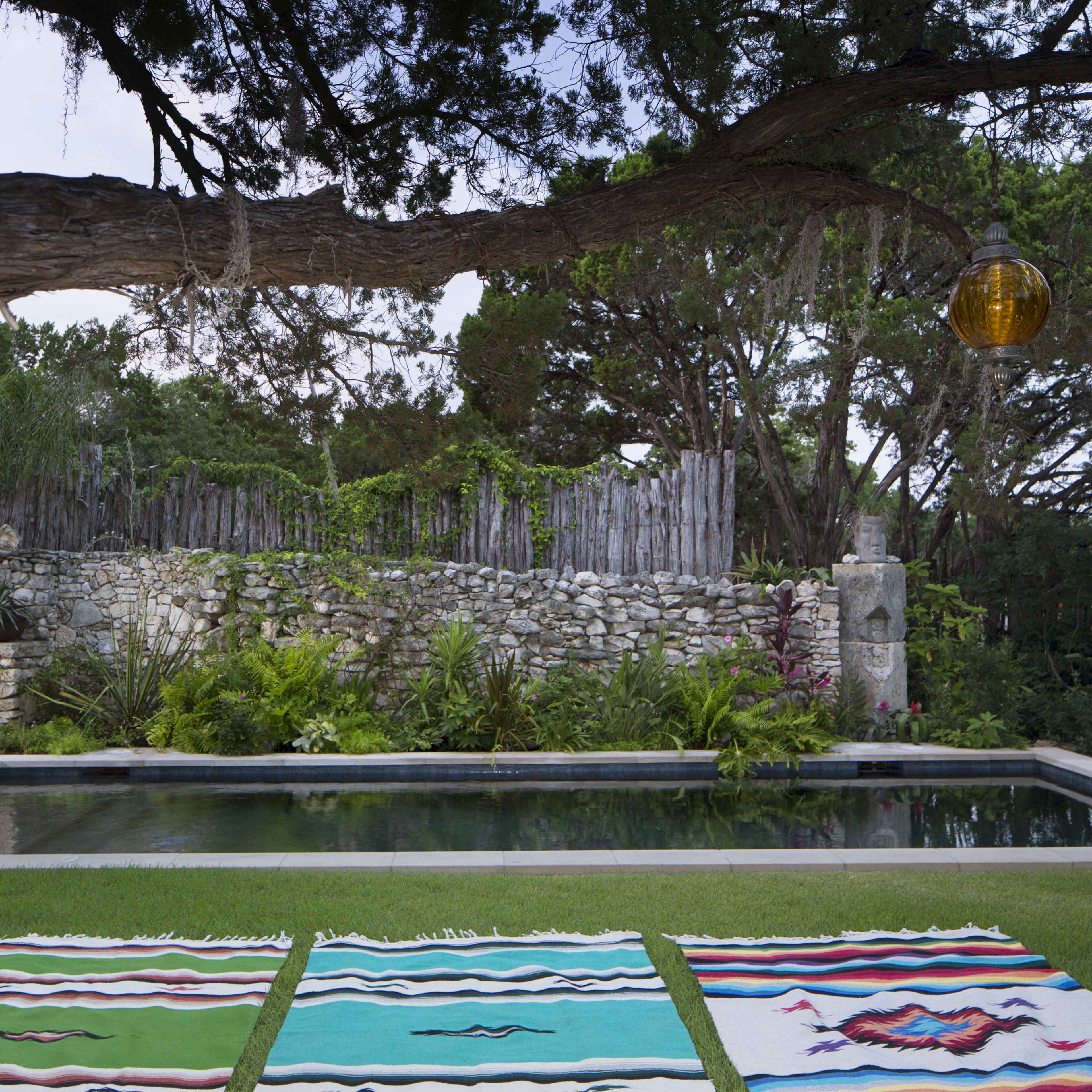 A serene outdoor scene with a rectangular swimming pool surrounded by lush greenery and a stone wall, with colorful woven blankets laid out on the grass in the foreground.