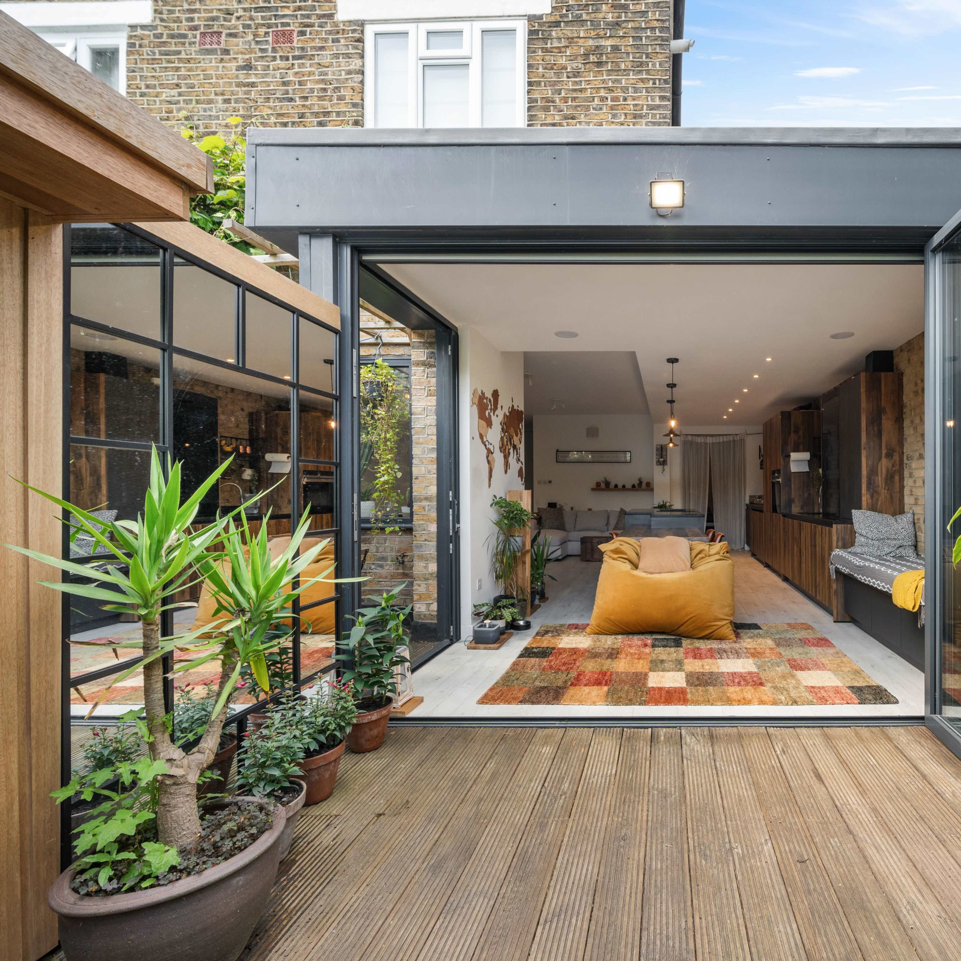 A modern living space with large glass doors open to a wooden deck surrounded by potted plants and a colorful rug inside.