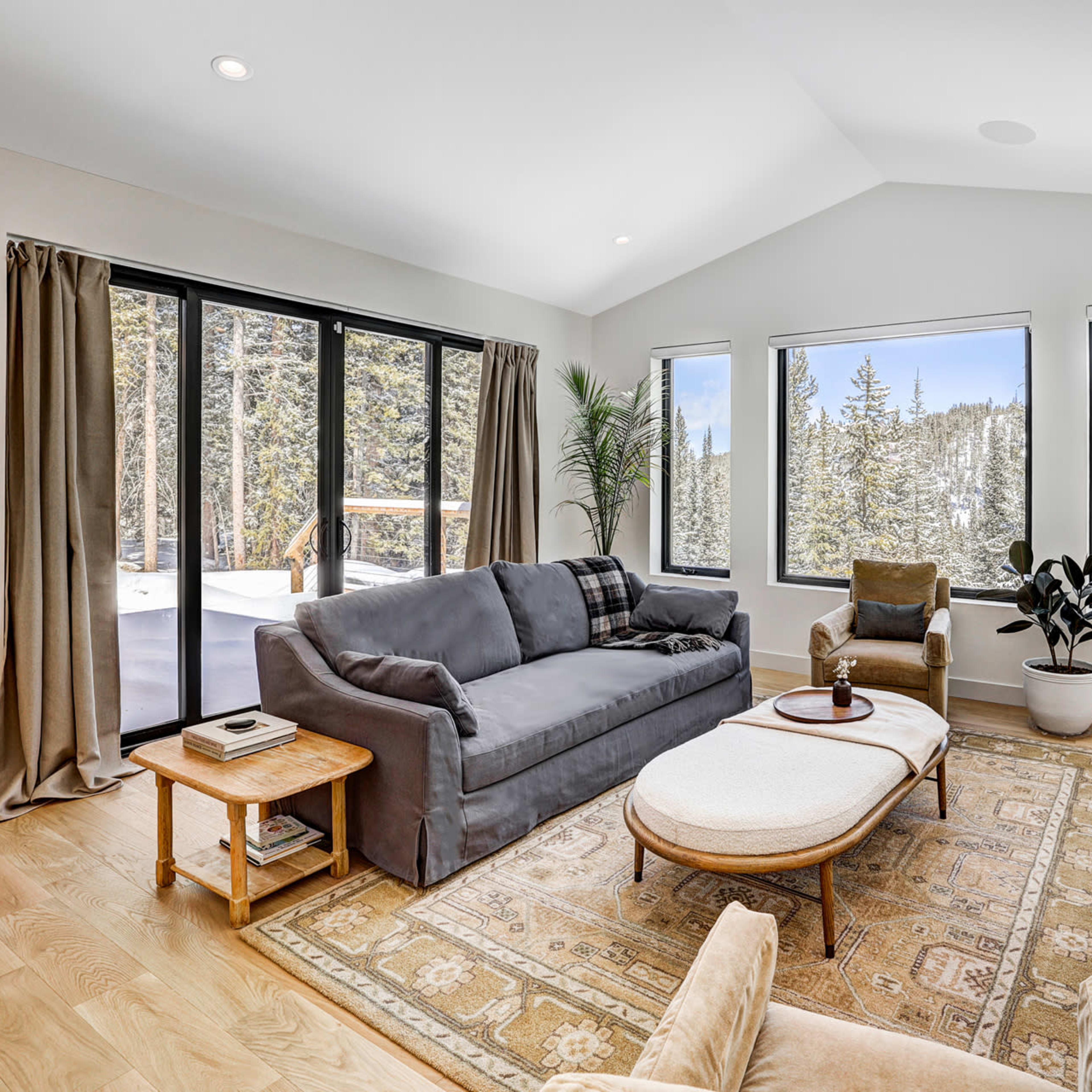 A modern living room features large windows overlooking a snowy landscape, with a sofa, armchair, and a coffee table on a patterned rug.