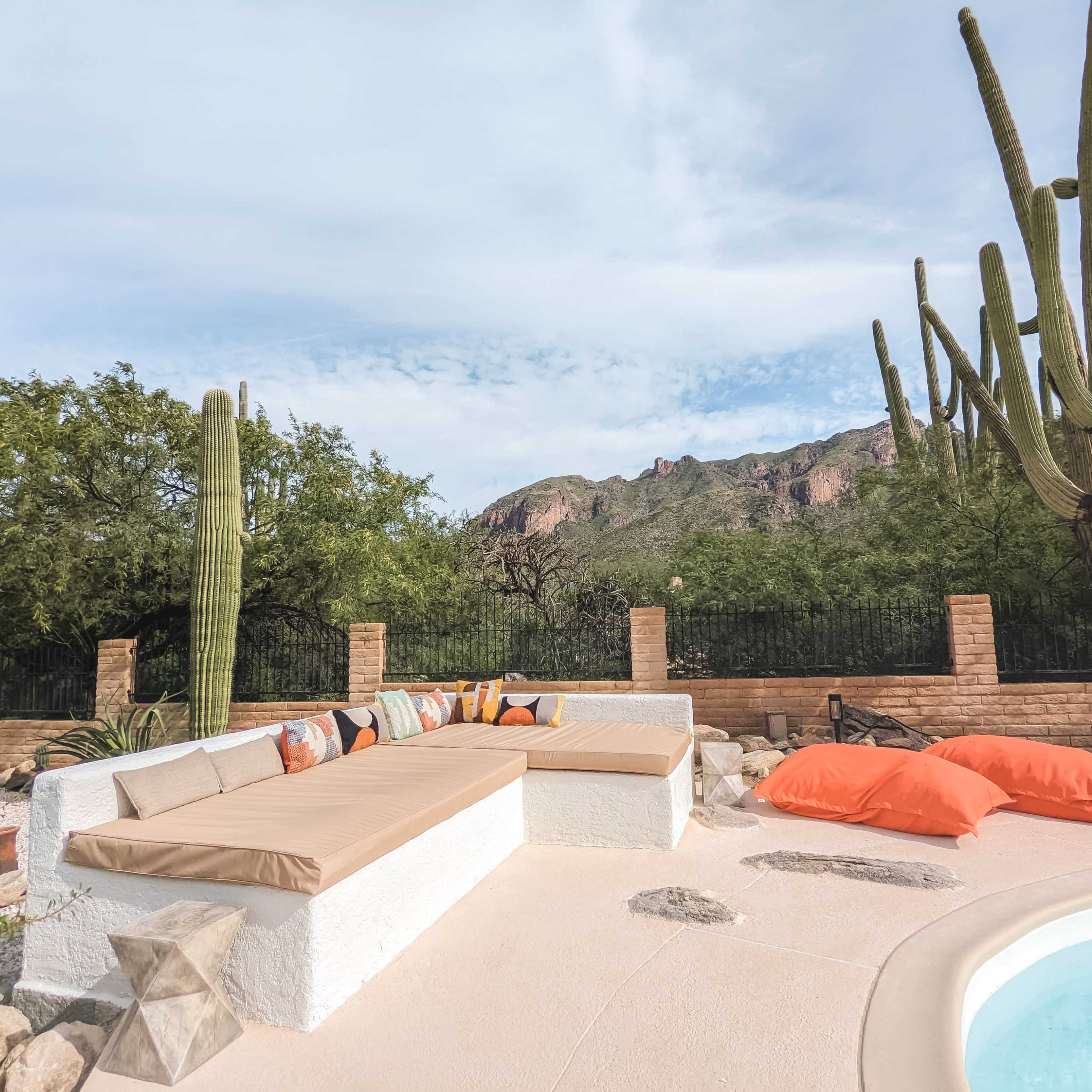 A poolside seating area with a cushioned bench and orange bean bags is set against a desert landscape featuring tall cacti and mountains in the background.