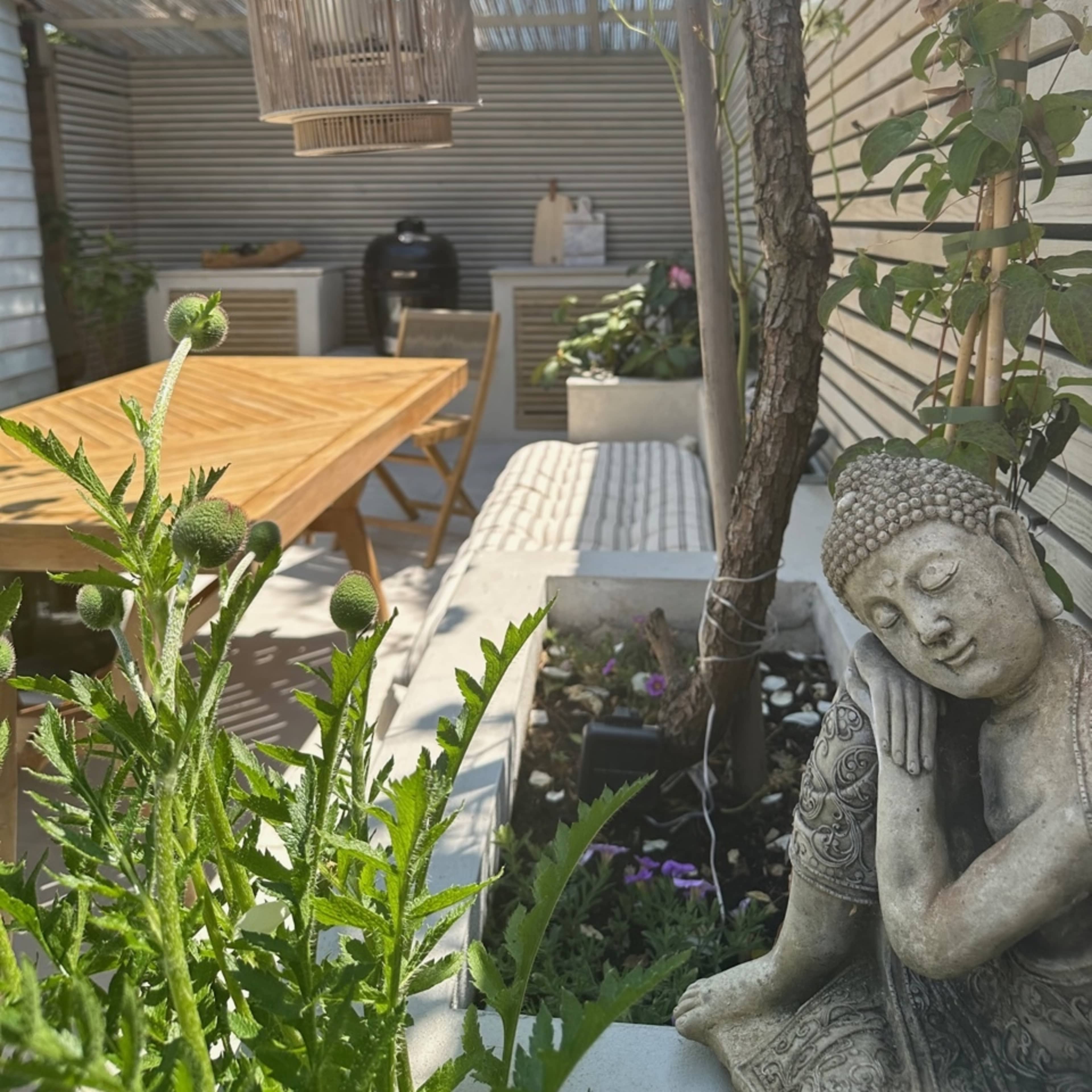 A serene garden scene with a wooden table, a Buddha statue, and a decorative lantern hanging from a tree, surrounded by greenery and potted plants.