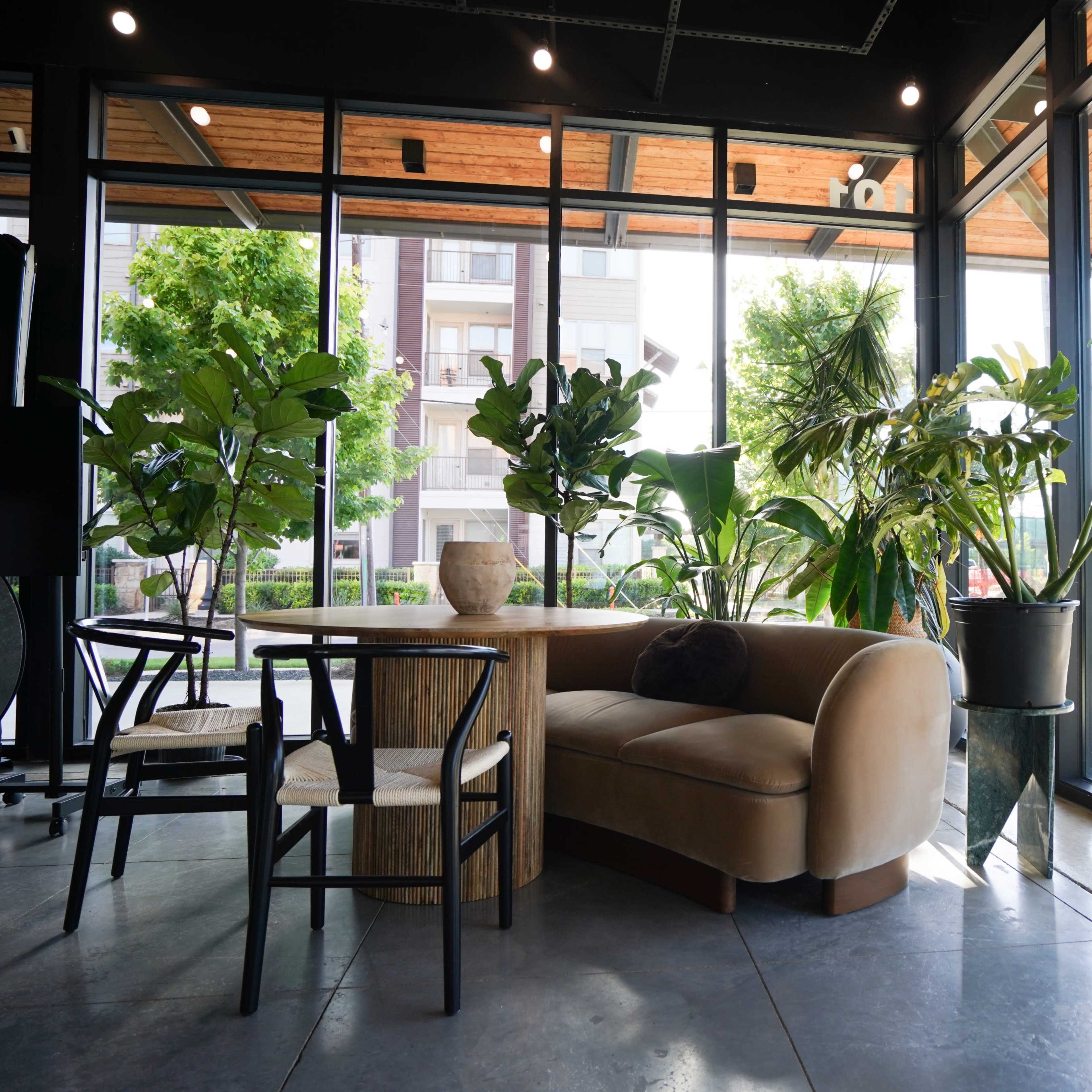 A cozy seating area is set within a glass-walled space, featuring a curved sofa, a wooden table, and various potted plants.