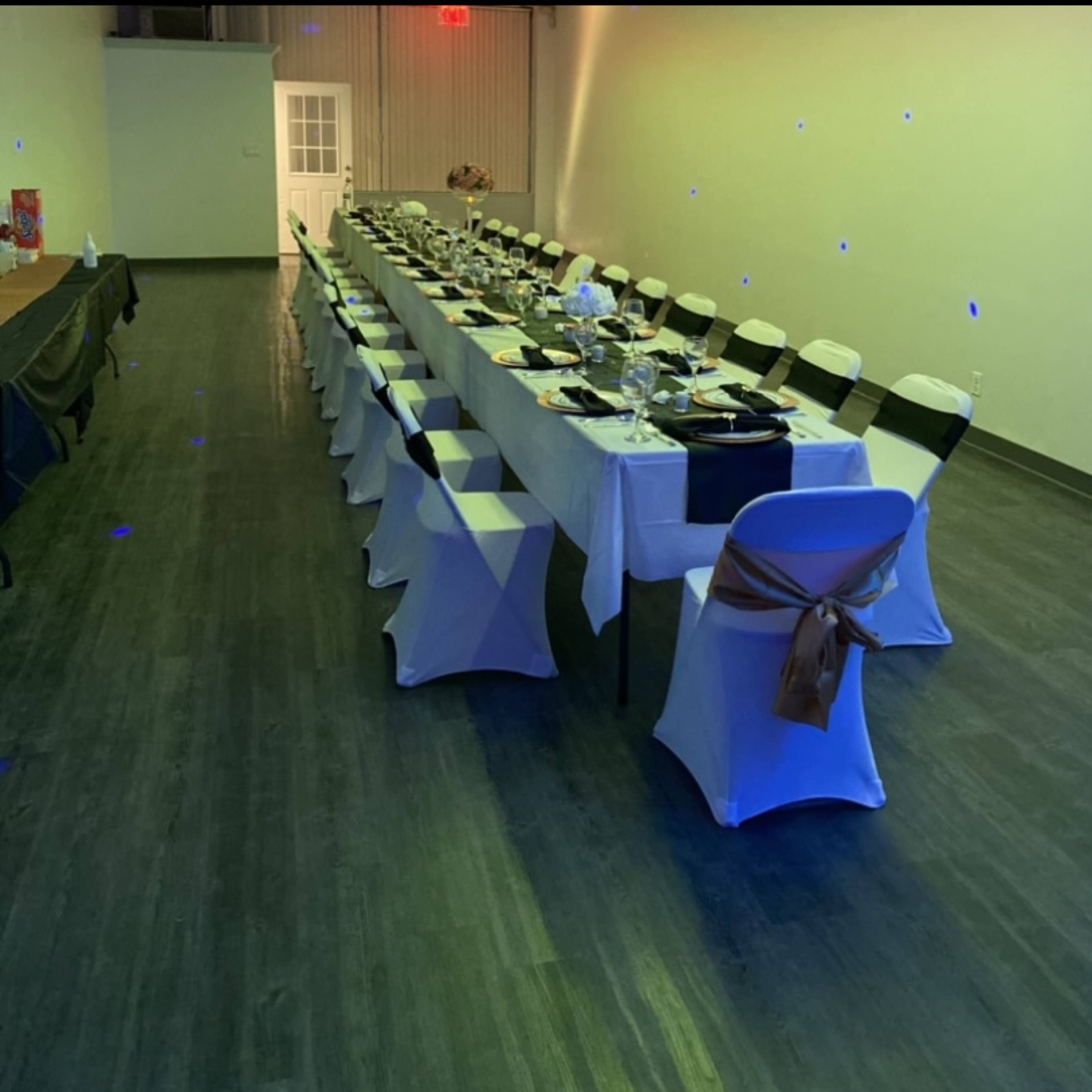 A long dining table set for a formal meal is arranged in a banquet hall with chairs covered in white and black fabric.