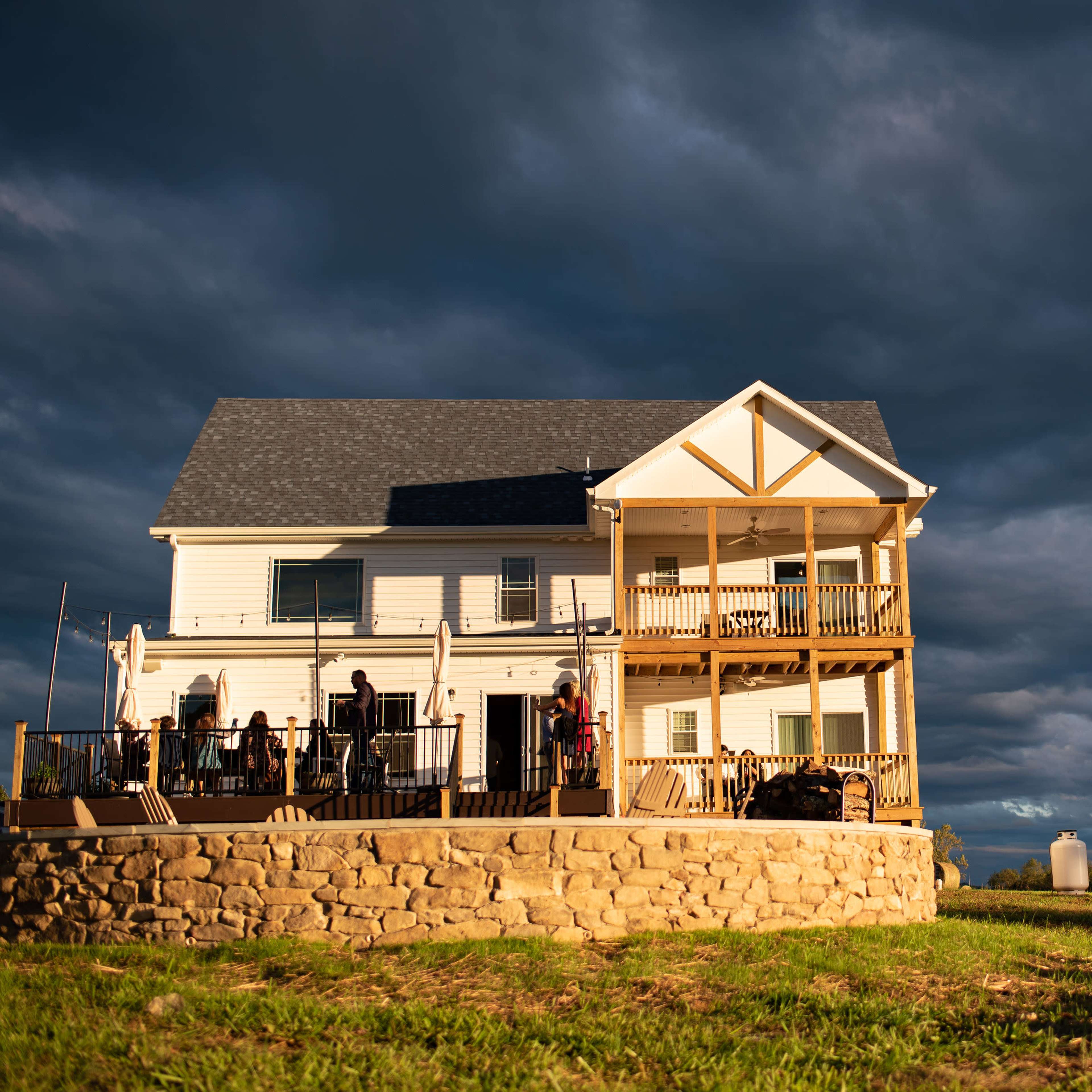 A two-story house with multiple balconies stands on a grassy hillside under a dramatic sky.