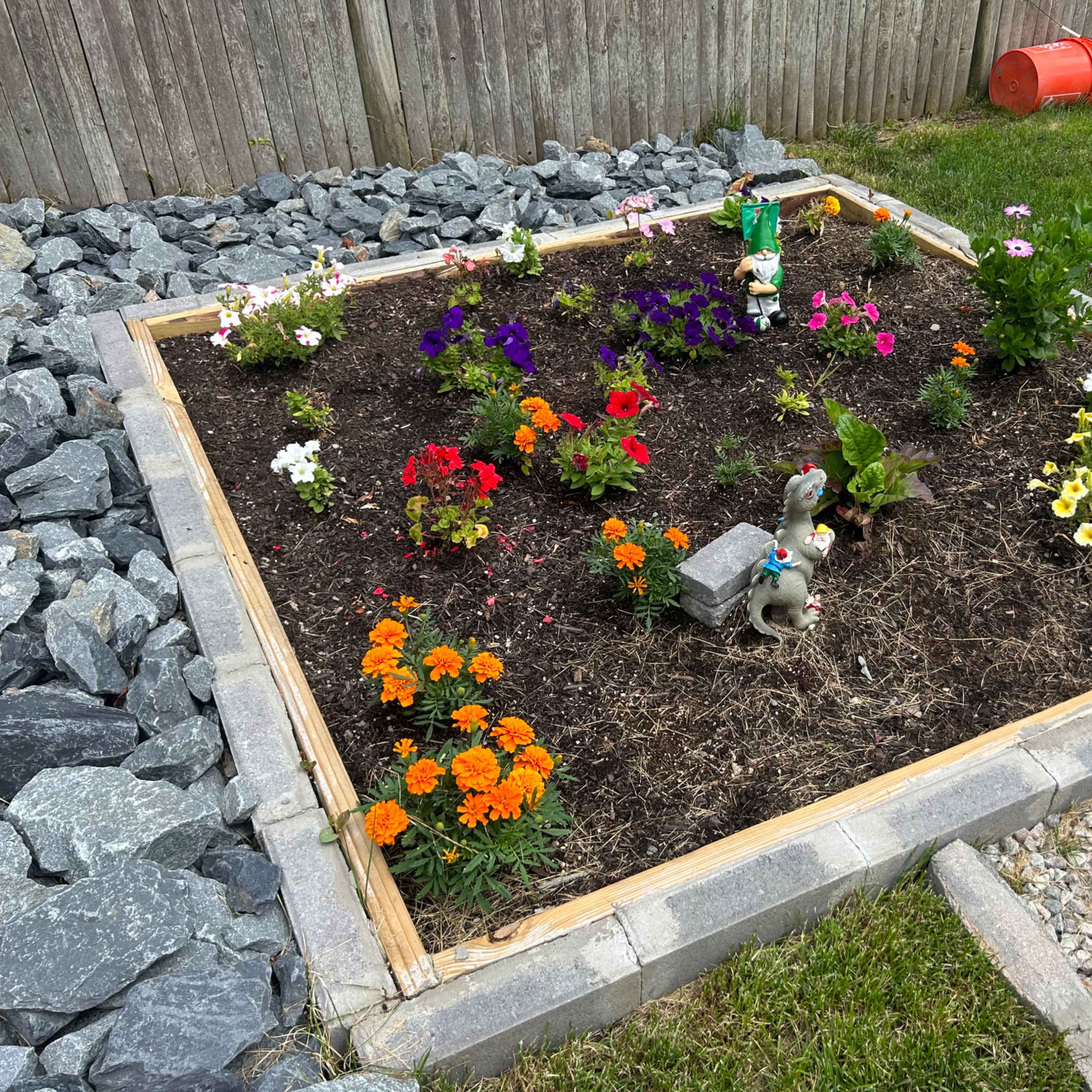 A flower garden is framed by wooden borders and contains various colorful flowers, a small statue, and decorative stones.