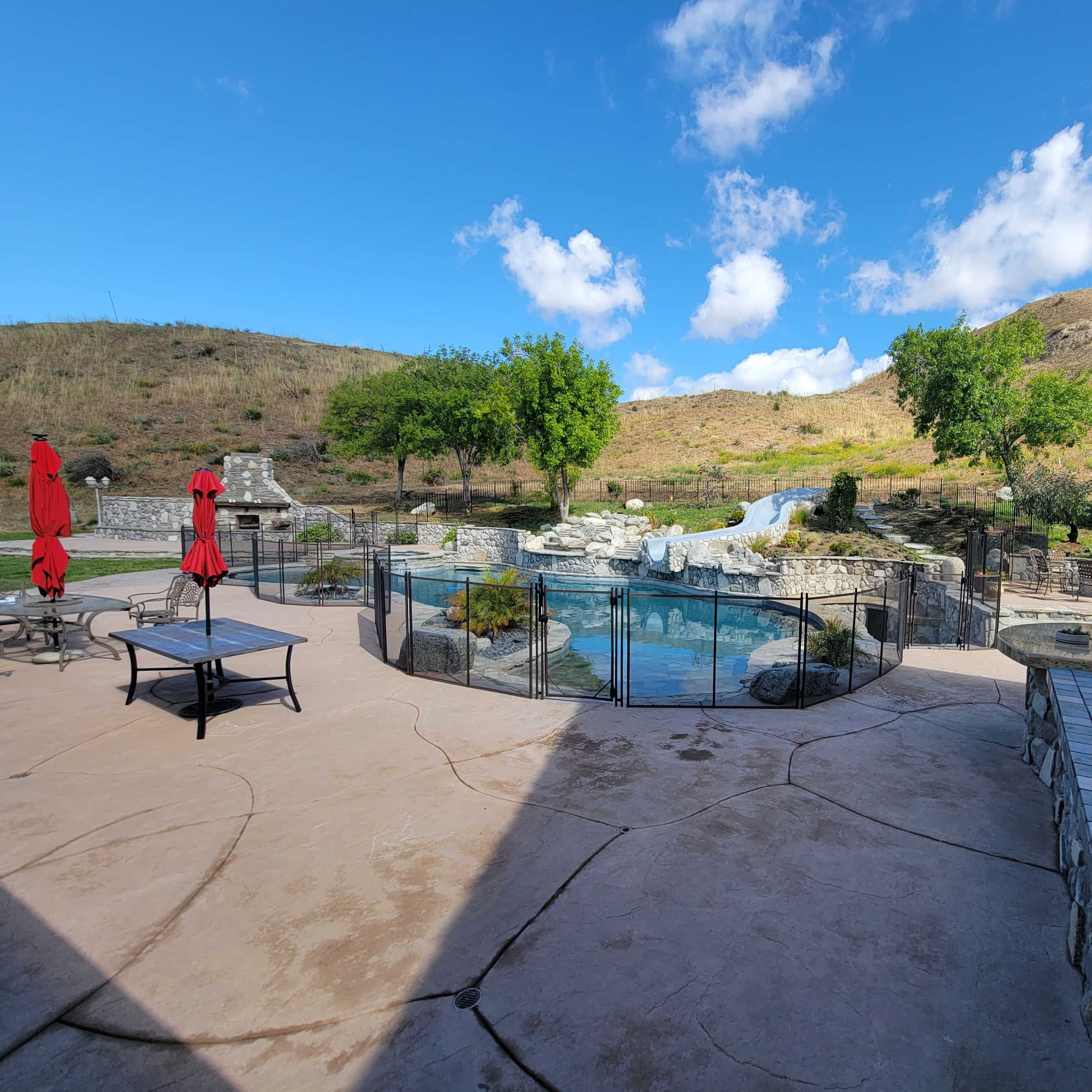 A landscaped outdoor area featuring a pool with a water slide, surrounded by greenery and stone structures, under a blue sky with few clouds.