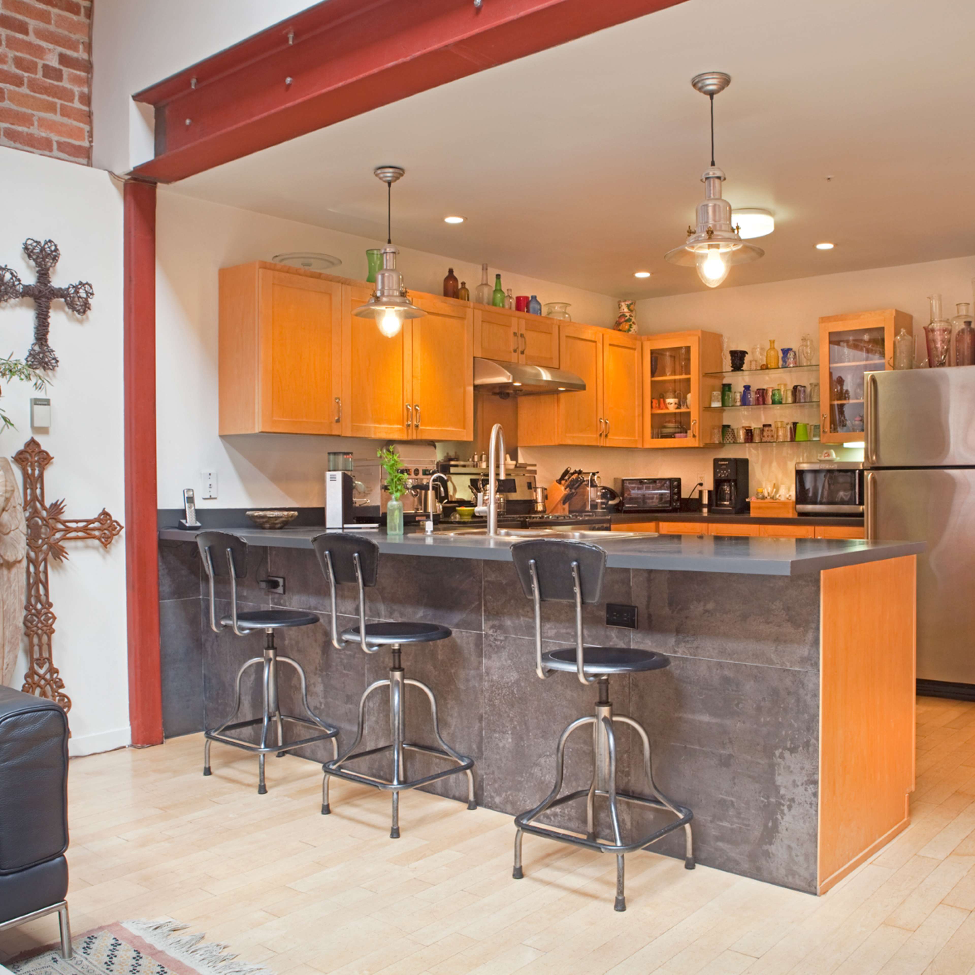 A modern kitchen with wooden cabinets, a stainless steel refrigerator, and a large island with bar stools, surrounded by a brick wall and decorative elements.