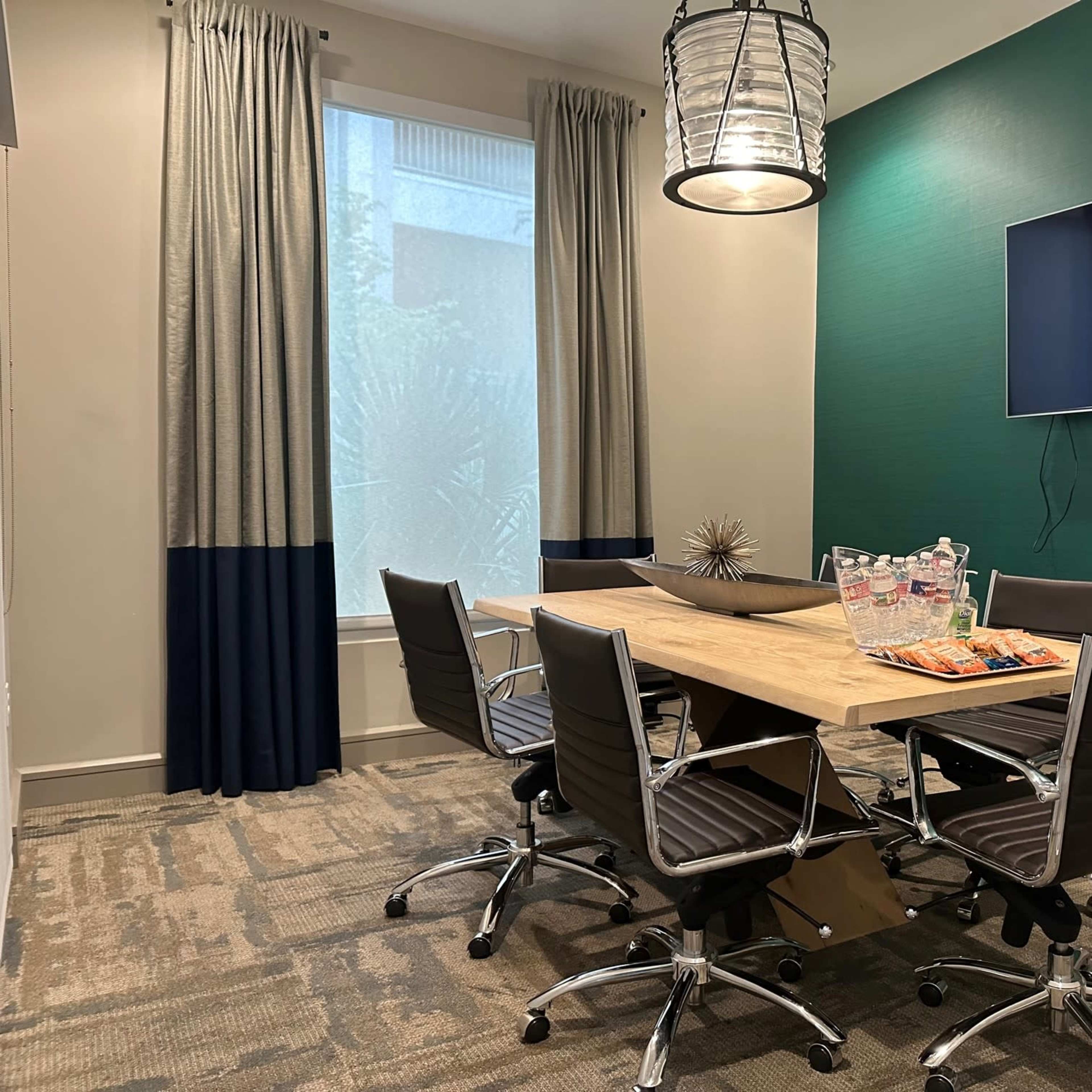 A conference room features a wooden table surrounded by six chairs, a television mounted on the wall, and curtains framing a window.