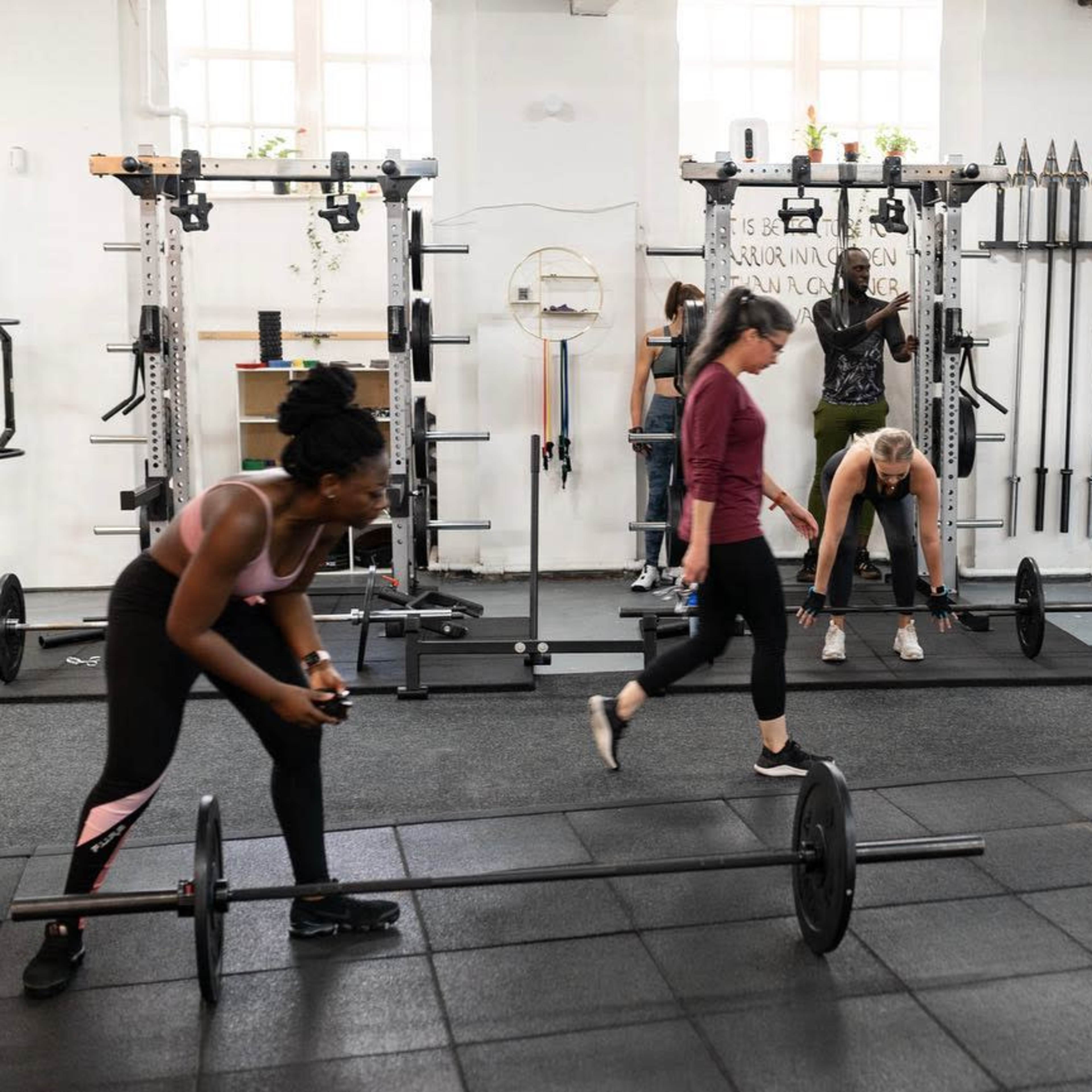 A group of women is working out in a gym with weightlifting equipment and exercise mats.