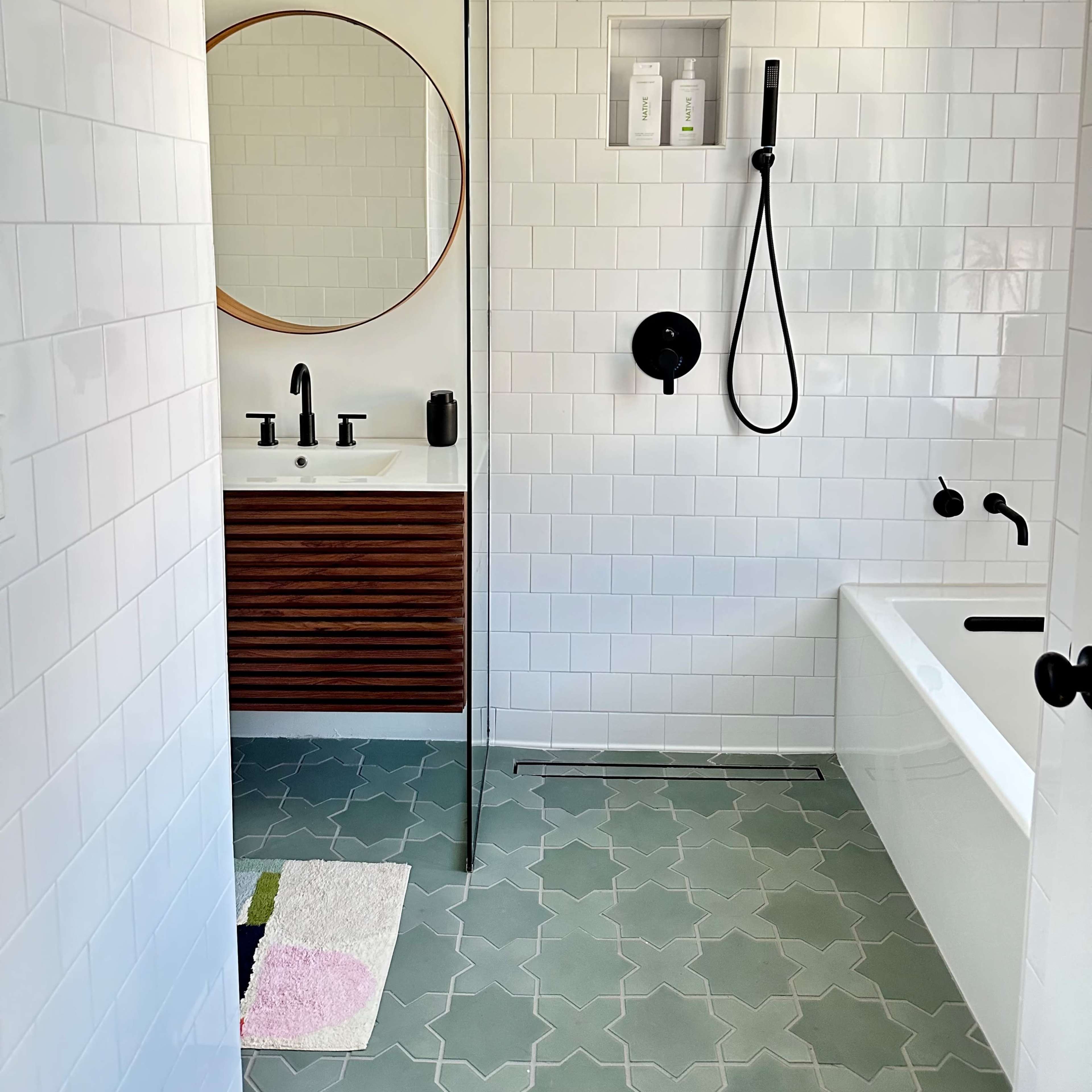 A modern bathroom features a walk-in shower with a rainfall showerhead, a freestanding bathtub, a round mirror above a sink with a wooden vanity, and patterned green tile flooring.