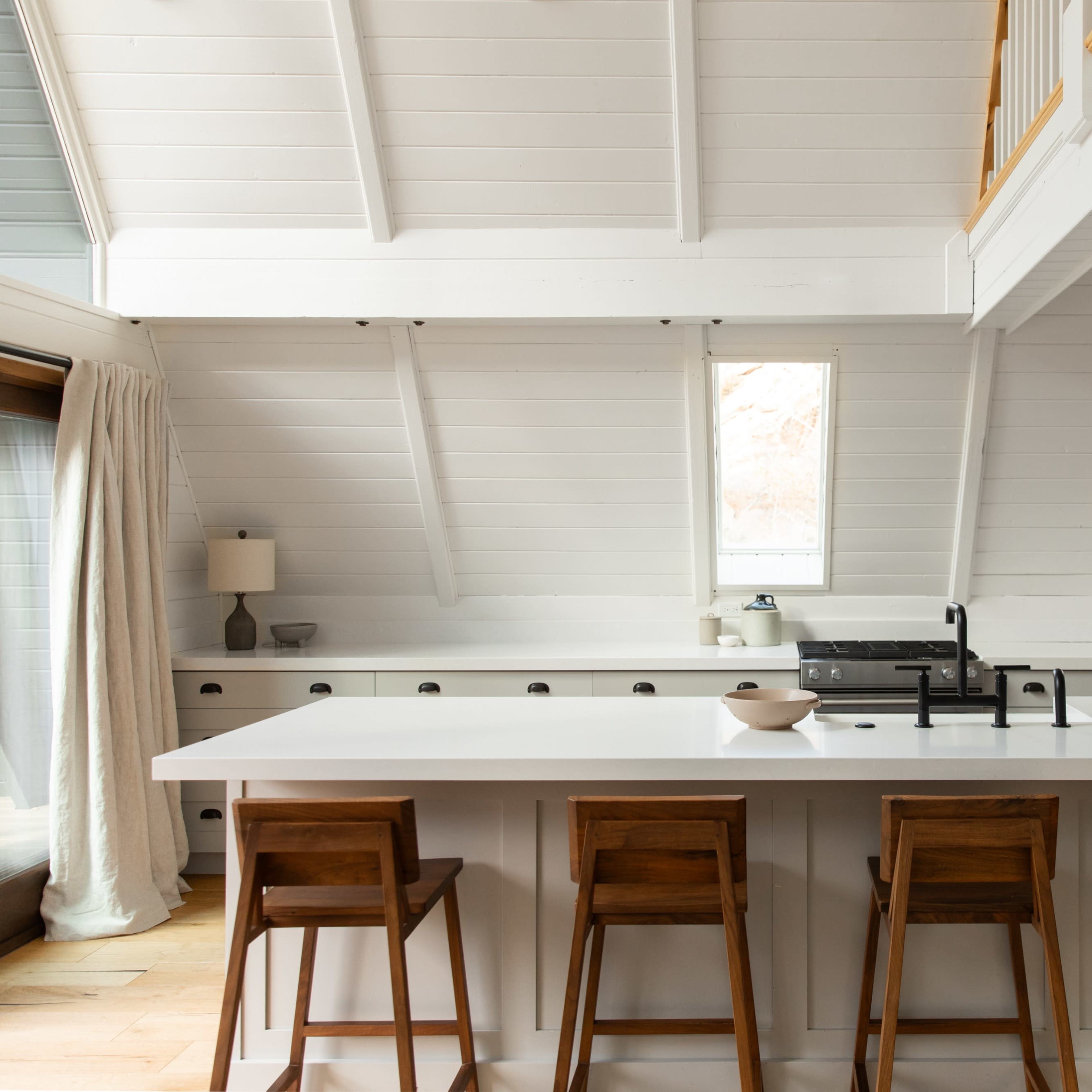 A modern kitchen with a white color scheme, featuring an island with four wooden bar stools and large windows allowing natural light.
