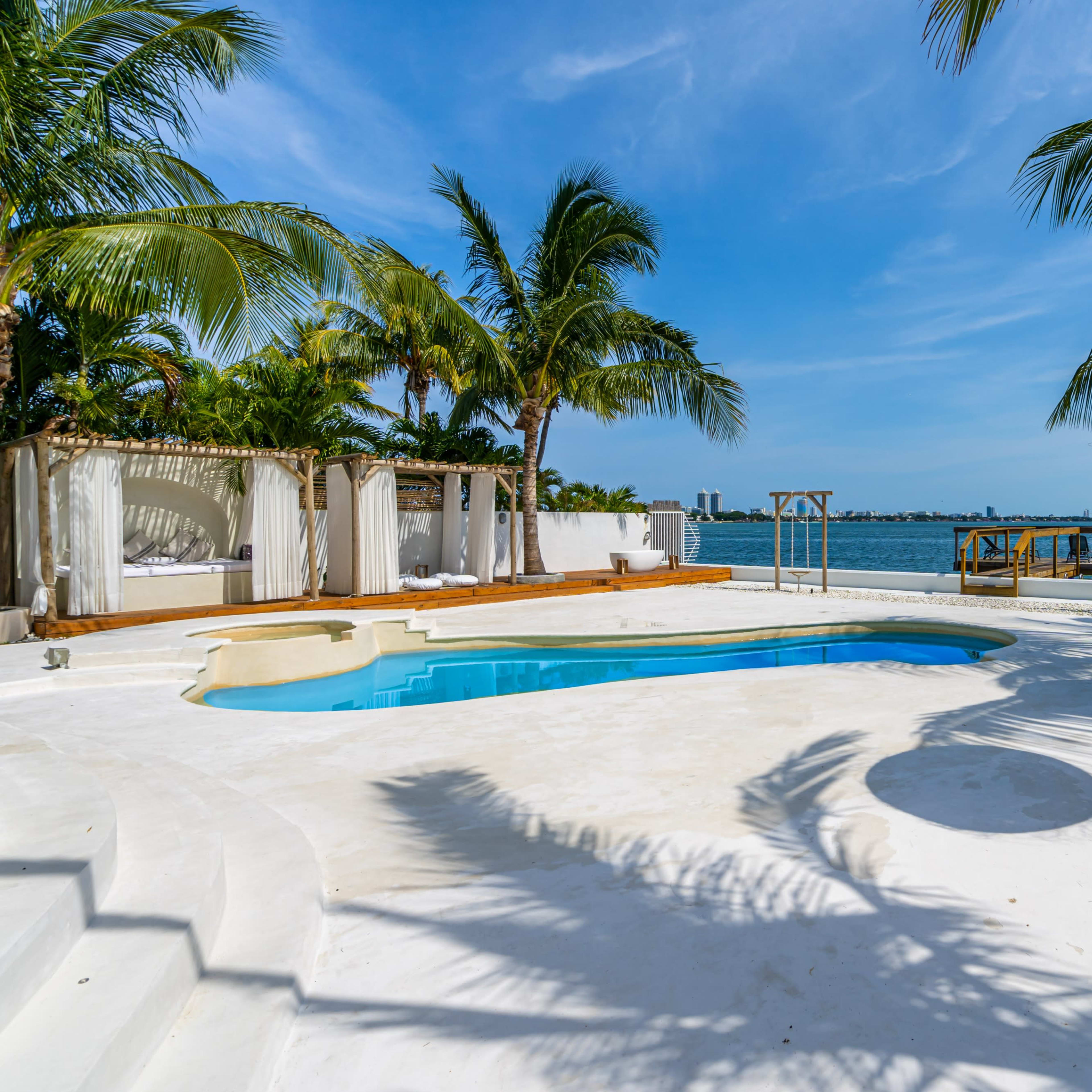 A poolside area with a swimming pool, palm trees, and a view of the water in the background.