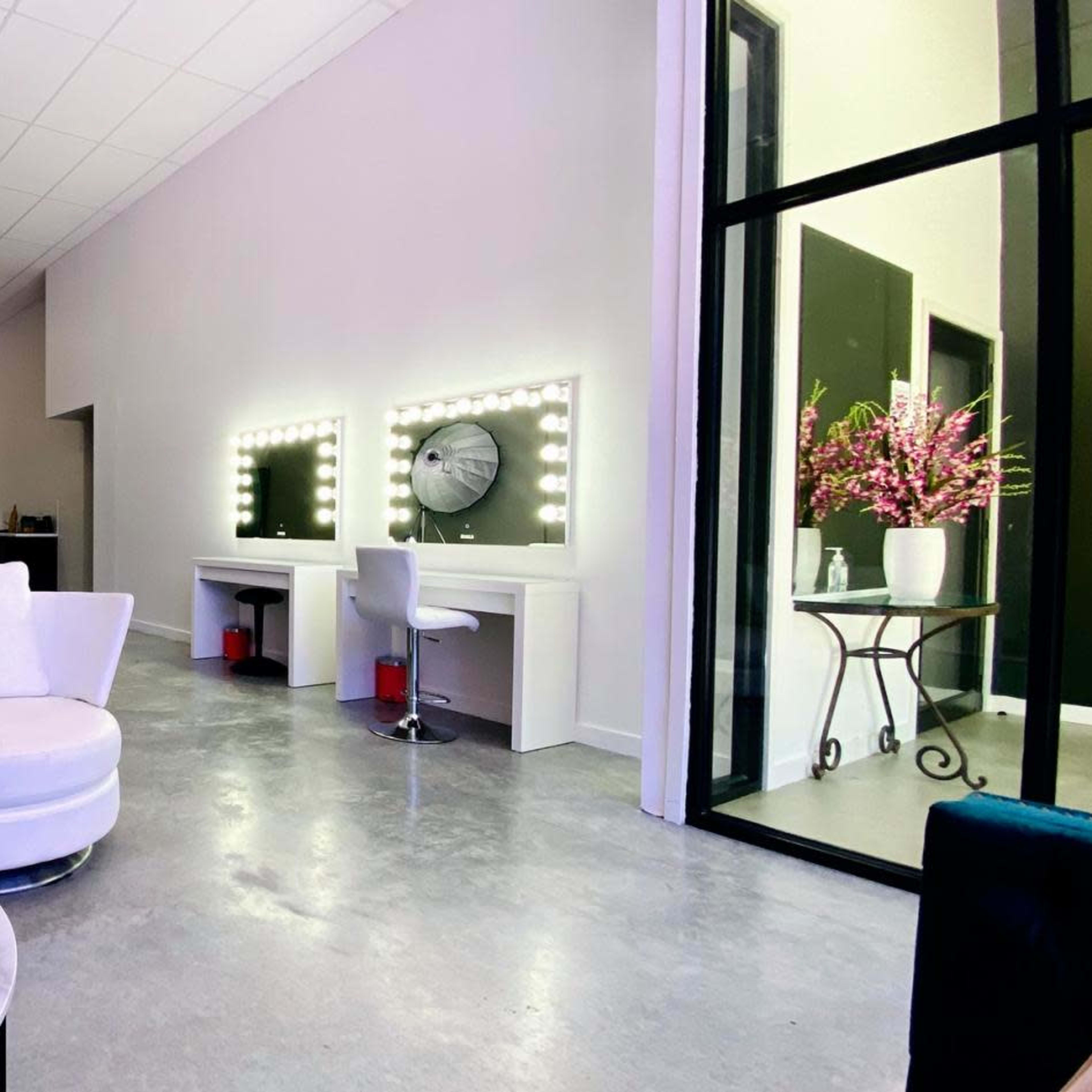 A modern interior of a cosmetic studio, featuring two illuminated mirrors, white circular chairs, a coffee table, and a large lighting setup in the background.