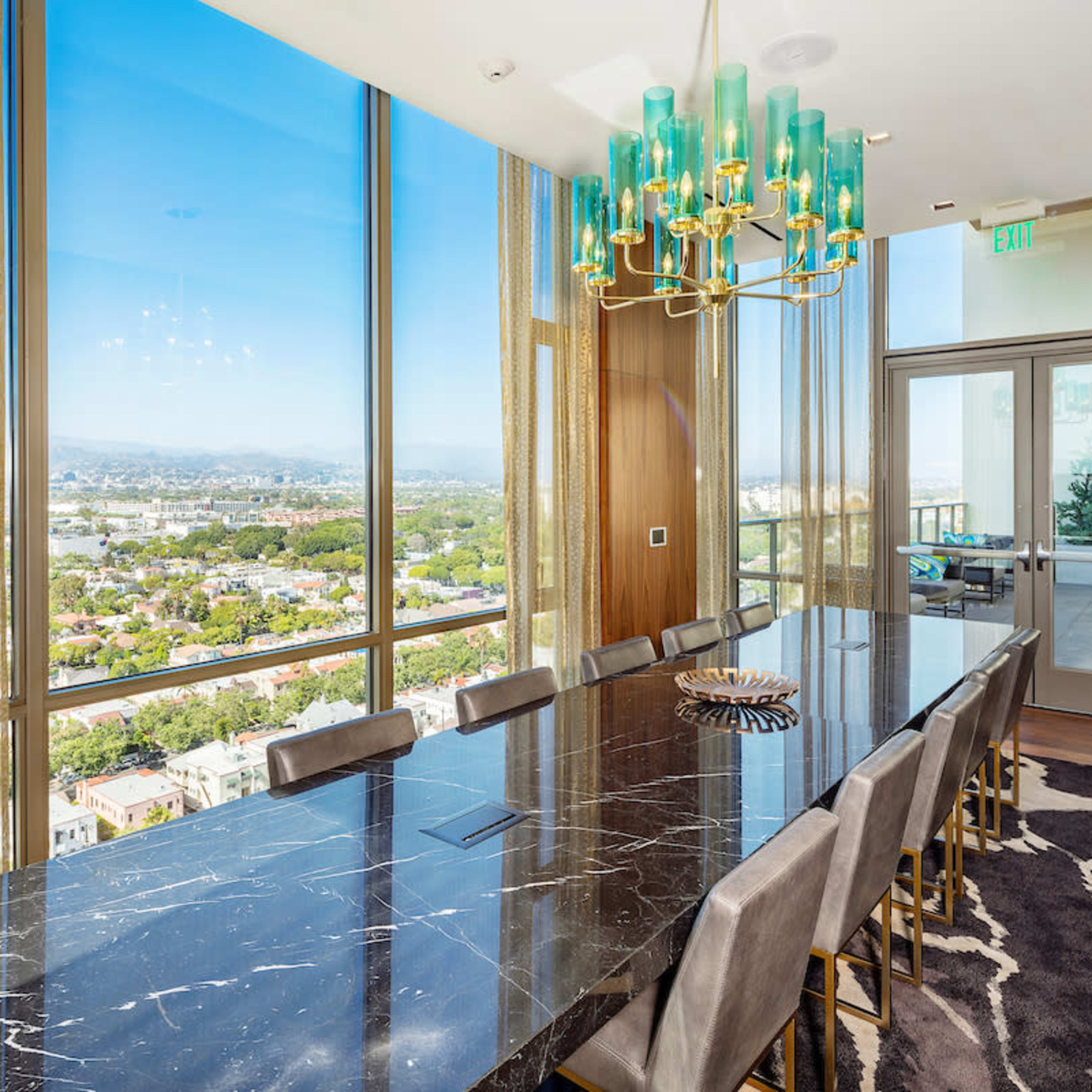 A modern dining room featuring a long black marble table surrounded by gray chairs, with large windows offering a view of the city and mountains.