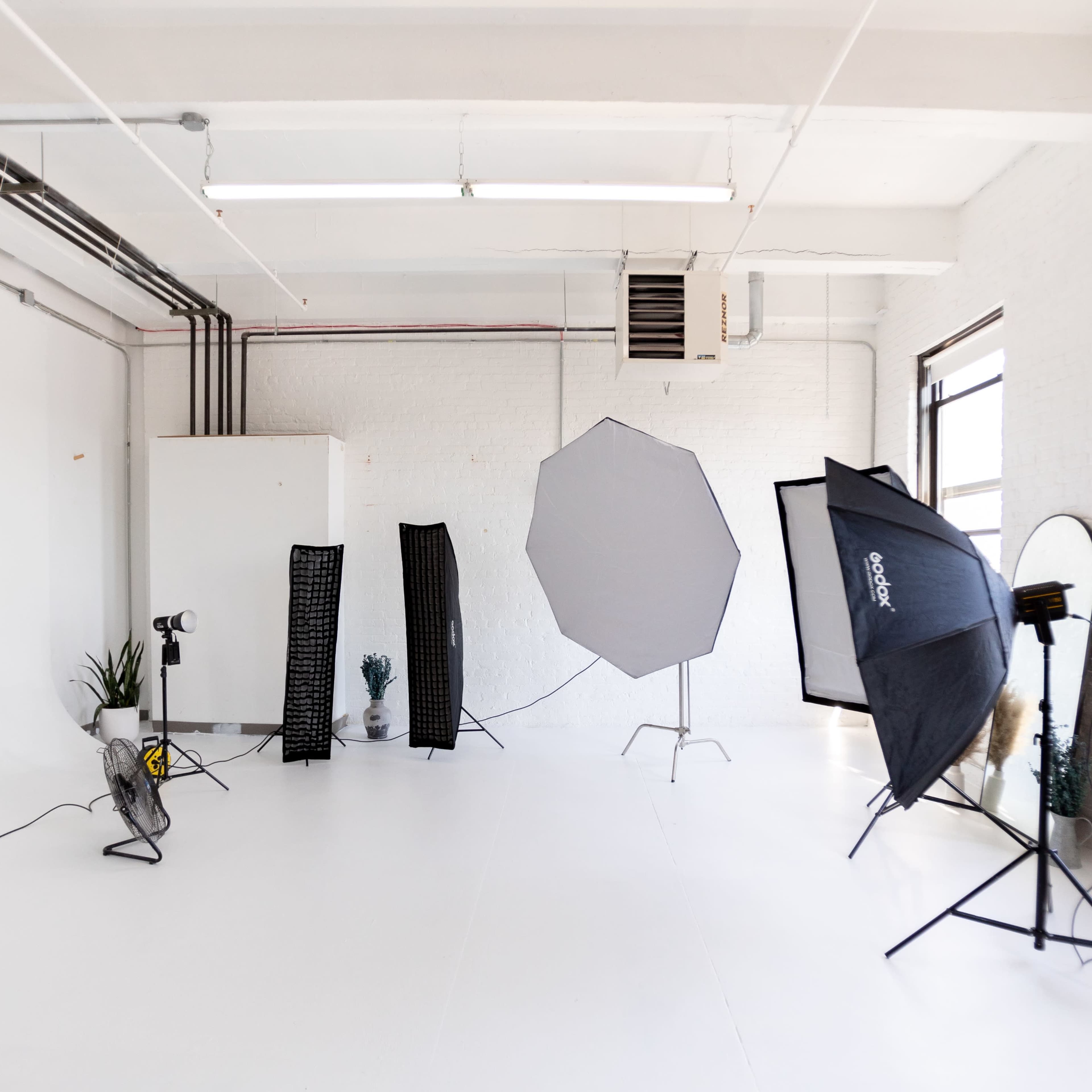 A photography studio with a clean, white backdrop, featuring various lighting equipment and props arranged around the space.