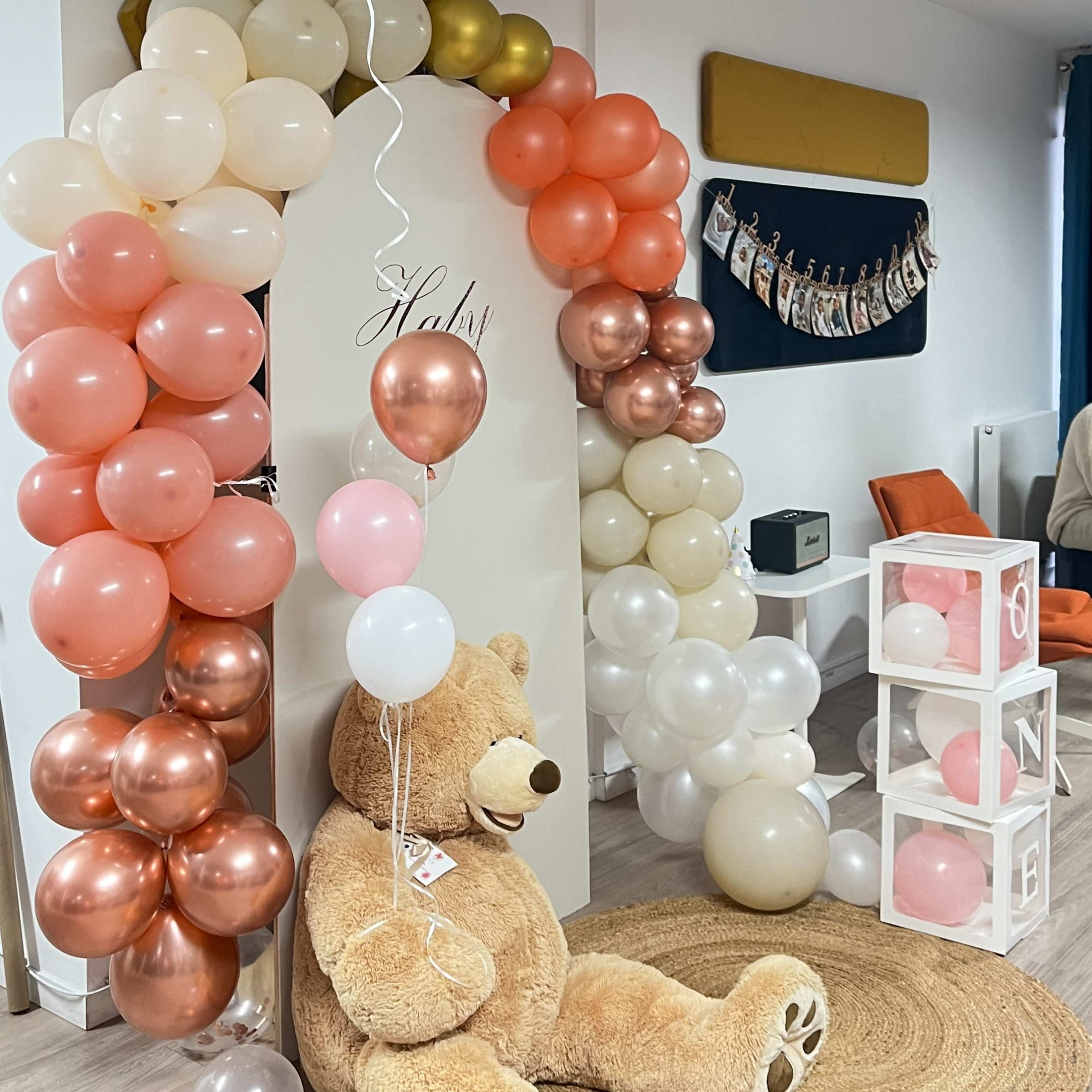 A large teddy bear sits on a rug under a decorative arch made of balloons in various colors, with a wall displaying photographs in the background.