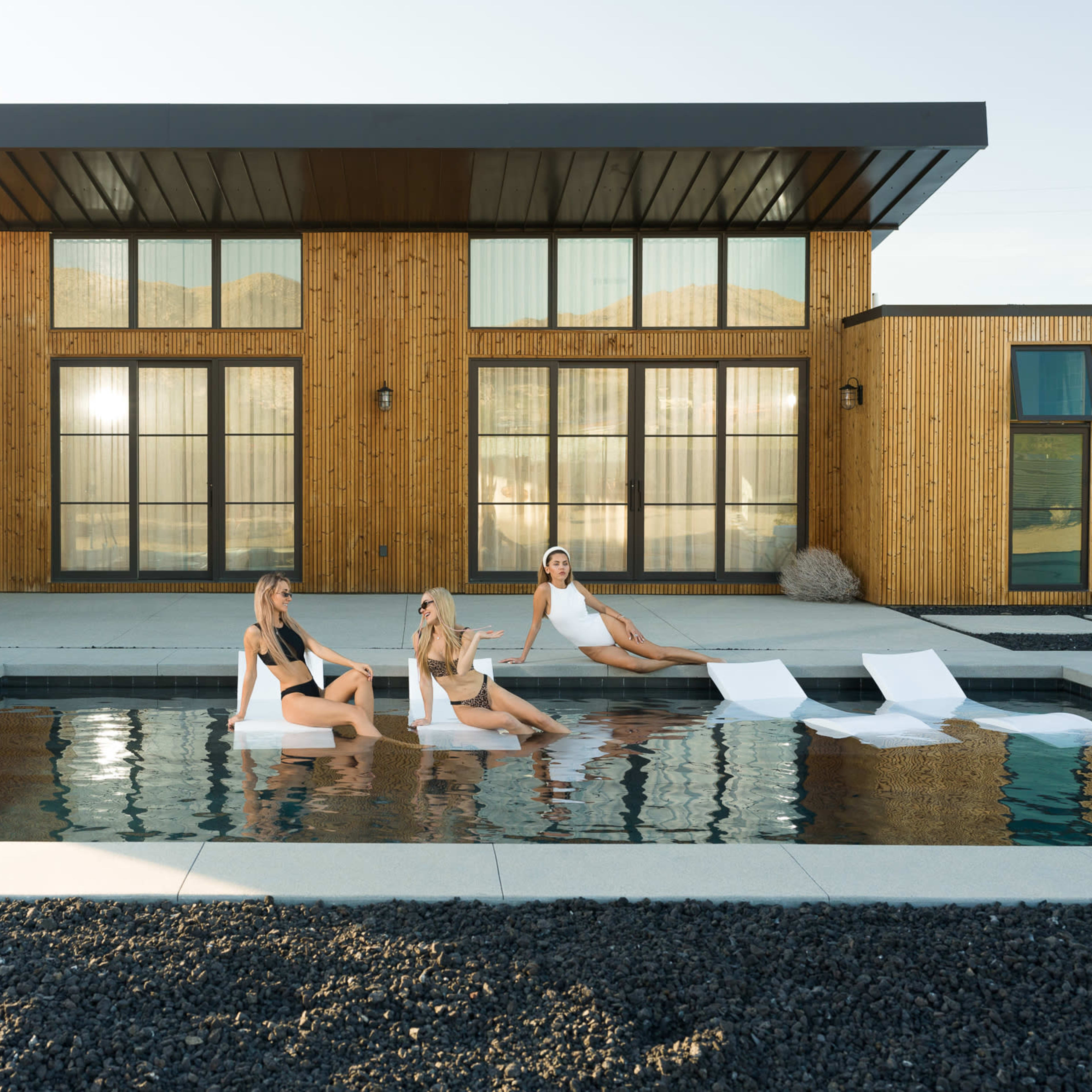 Three women relax on floating loungers in a modern pool in front of a stylish wooden-facade house.
