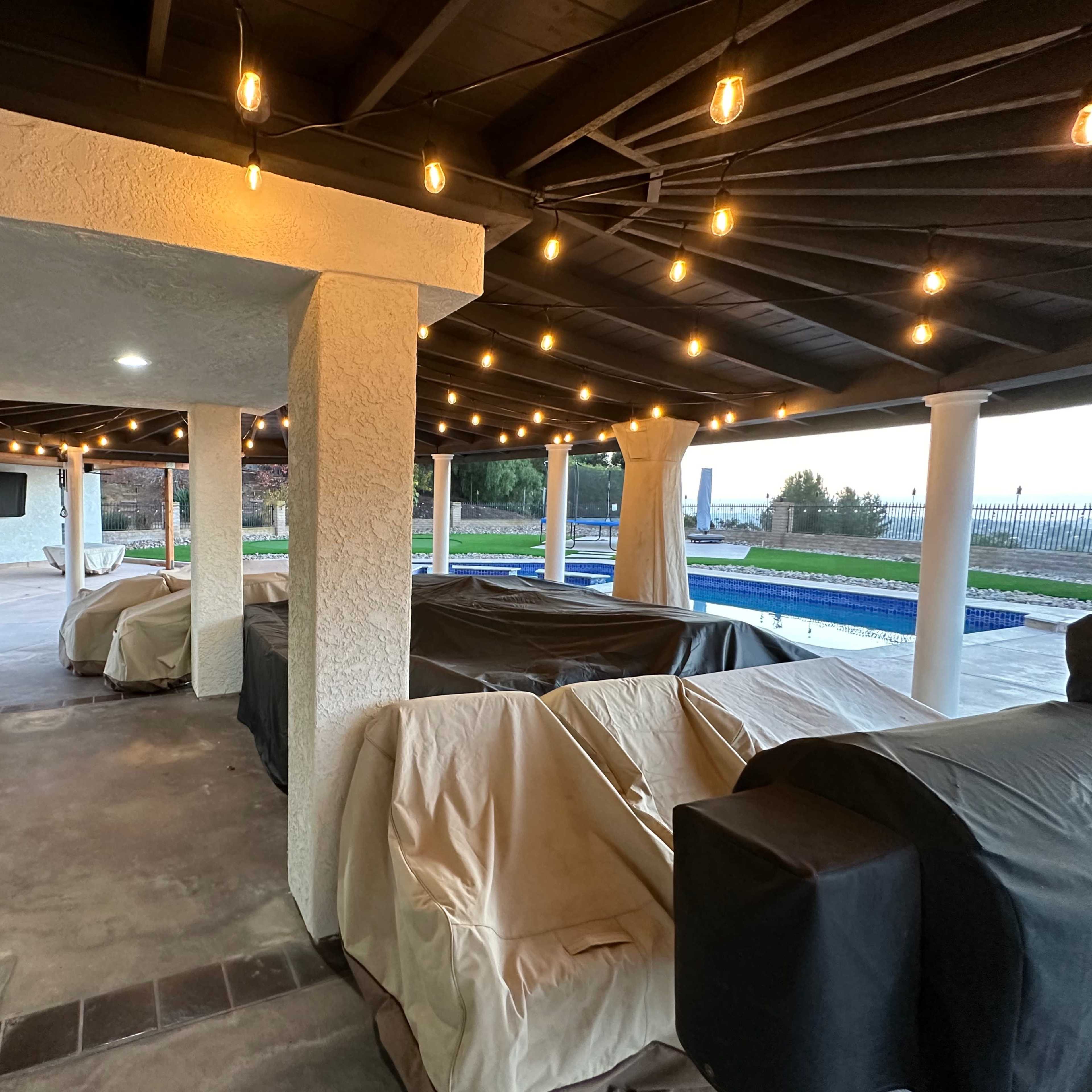 A covered outdoor patio area with string lights, furnishings covered with tarps, and a view of a pool beyond.