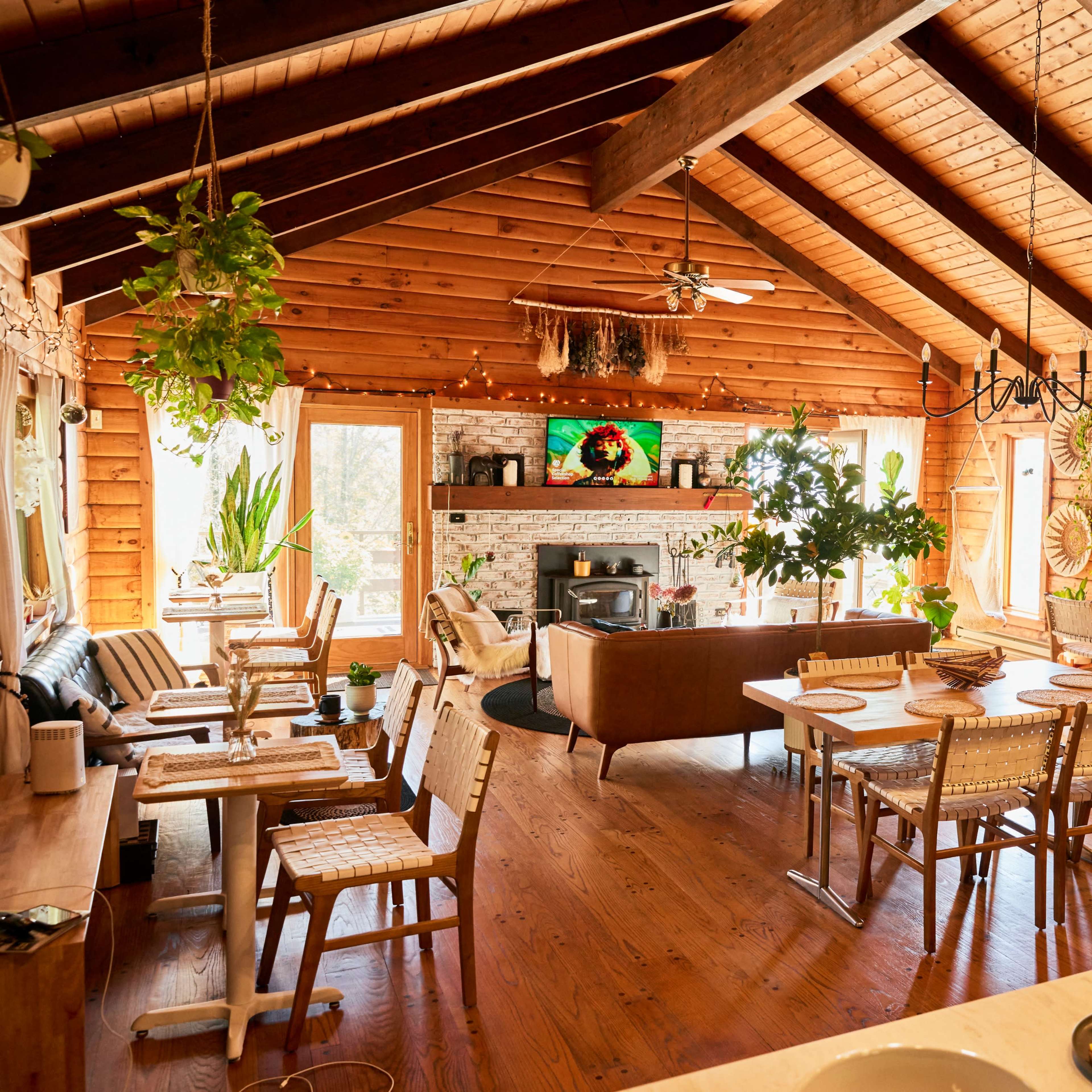 A spacious wooden interior features a dining area with several chairs and tables, a living space with a fireplace, and large windows allowing natural light.
