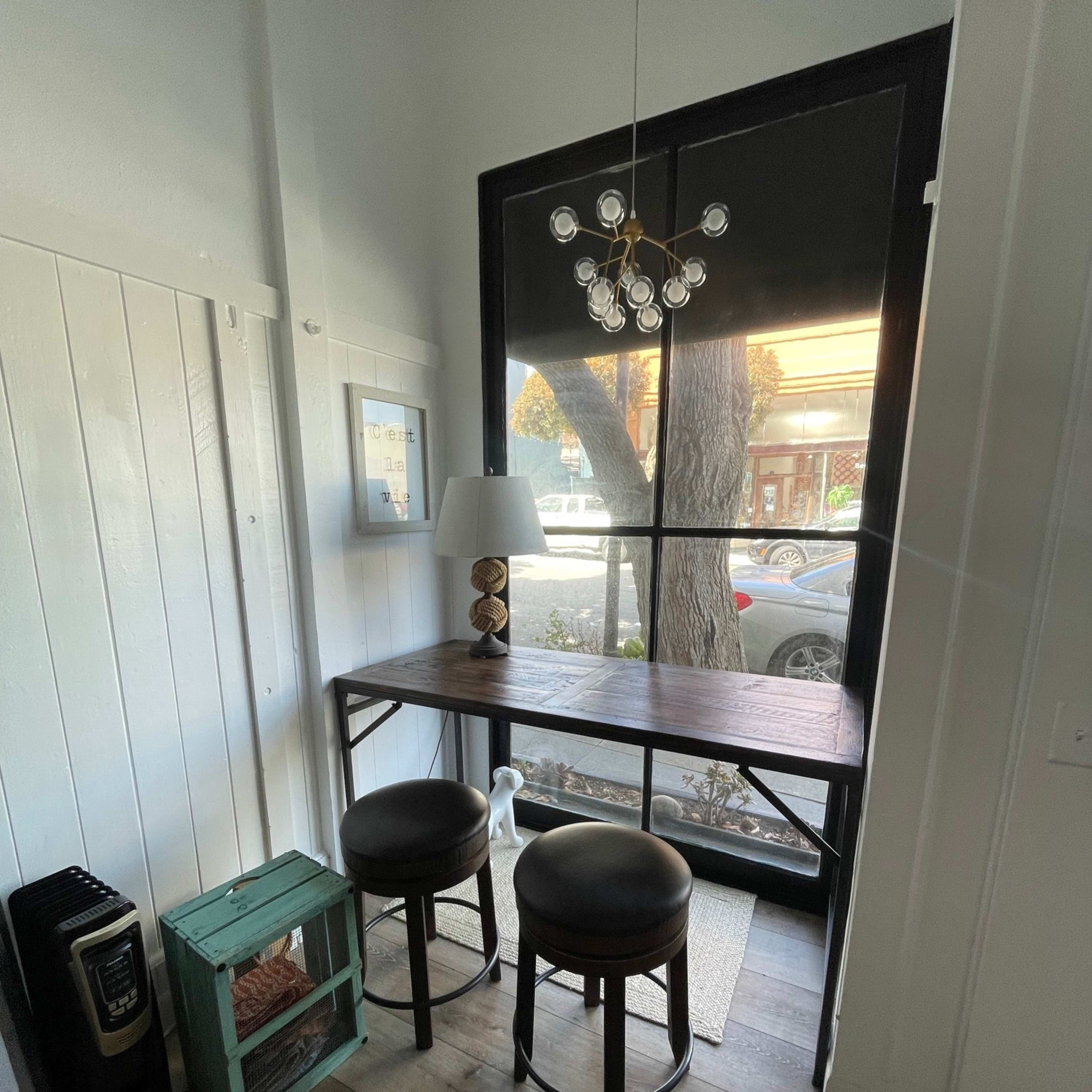 A small corner nook features a wooden table with two stools, a lamp, and a decorative box, all placed by a large window overlooking the street.