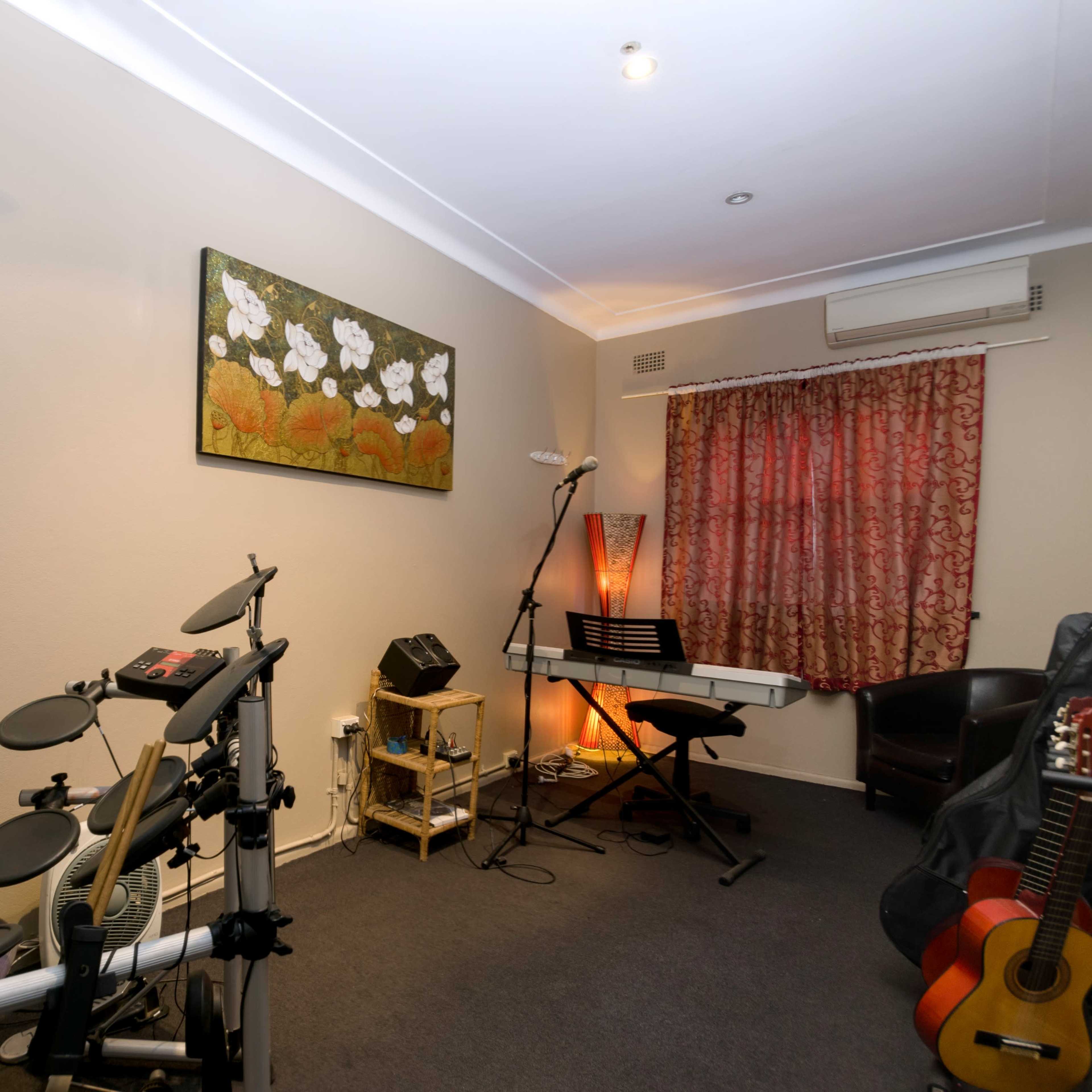 A room contains an electronic drum set, a keyboard on a stand, a chair, and various musical instruments against the wall, with a decorative painting and curtains.