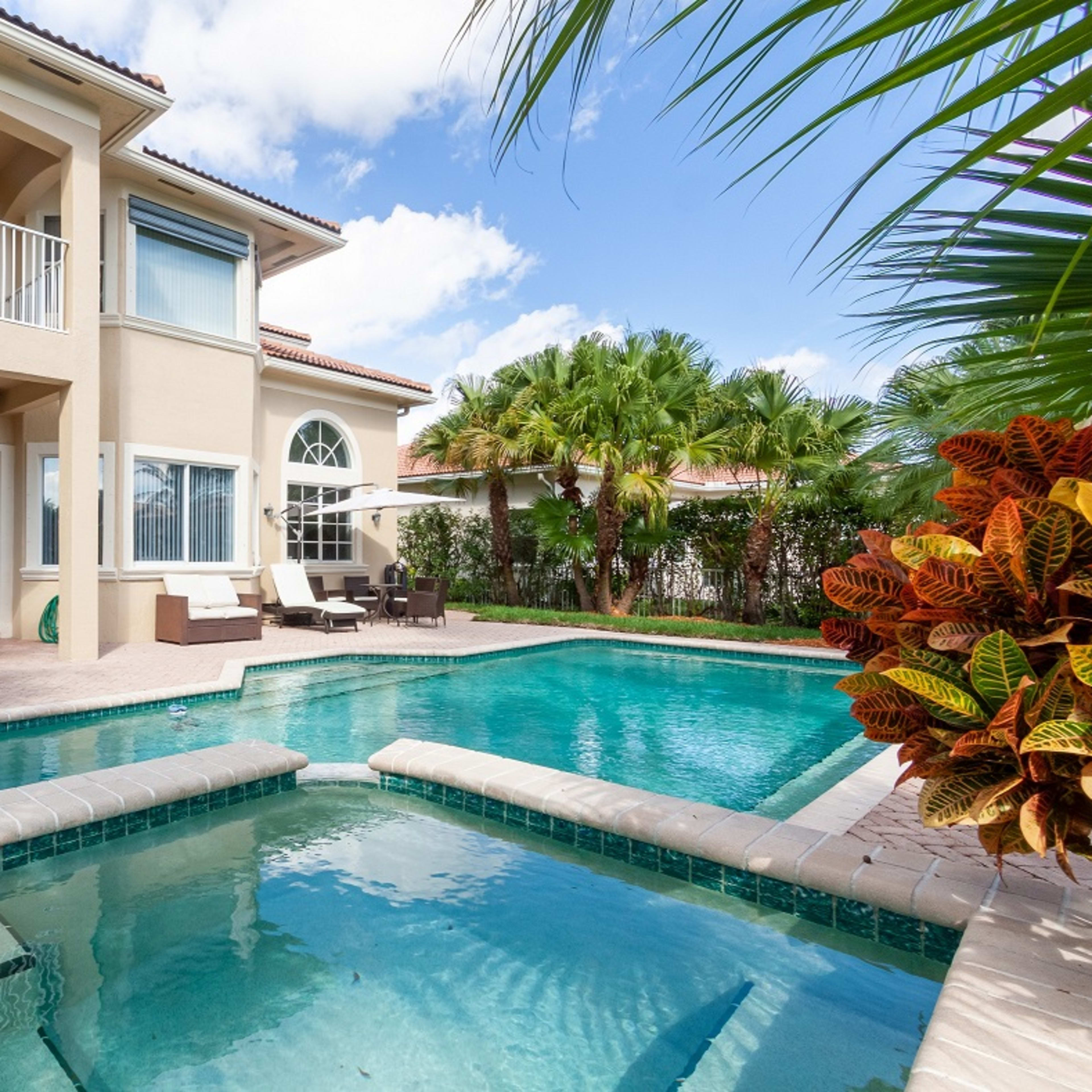A large two-story home with a swimming pool is surrounded by tropical landscaping and patio furniture.
