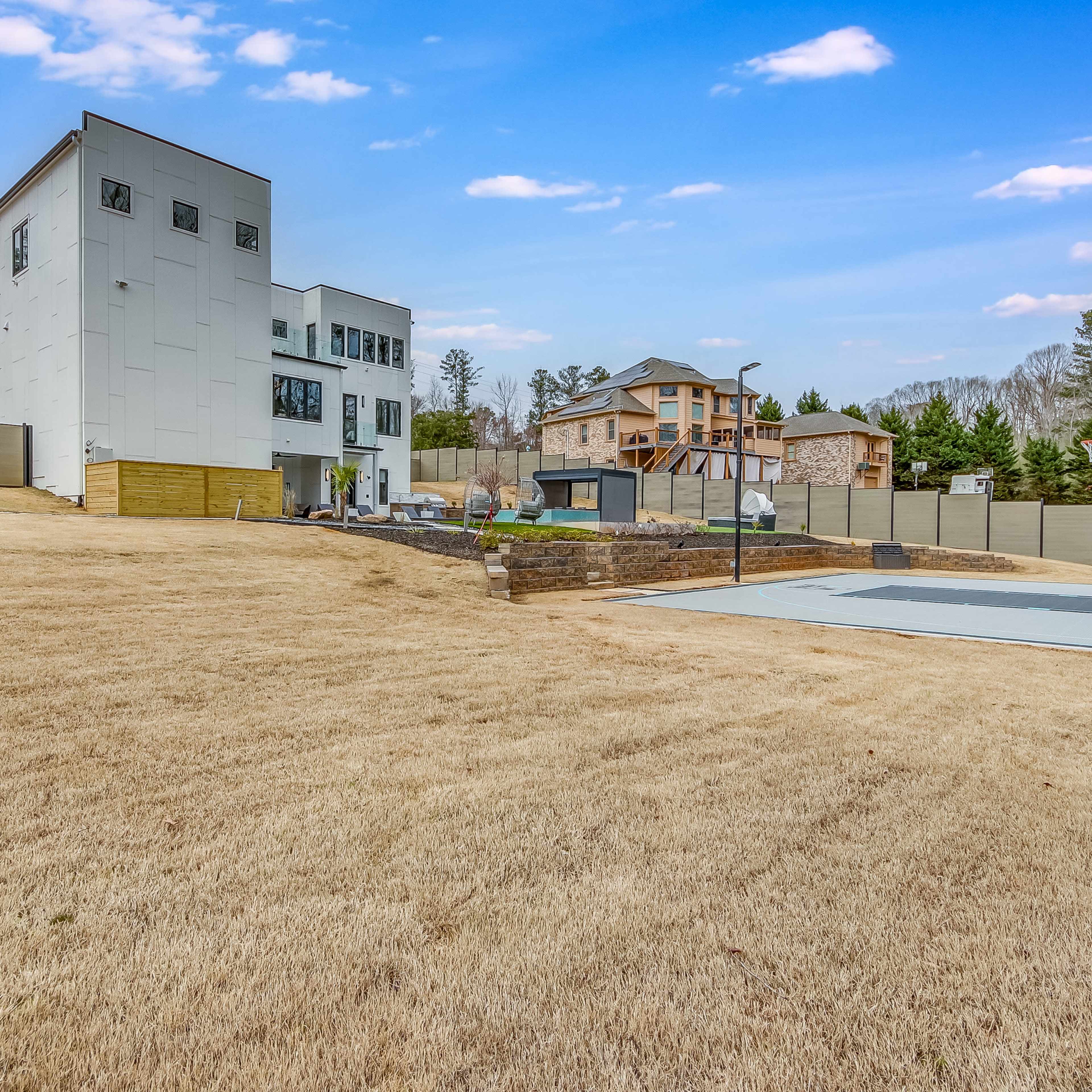 A multi-story modern building adjacent to a cleared area featuring a basketball court and surrounded by a wooden fence and other residential structures.