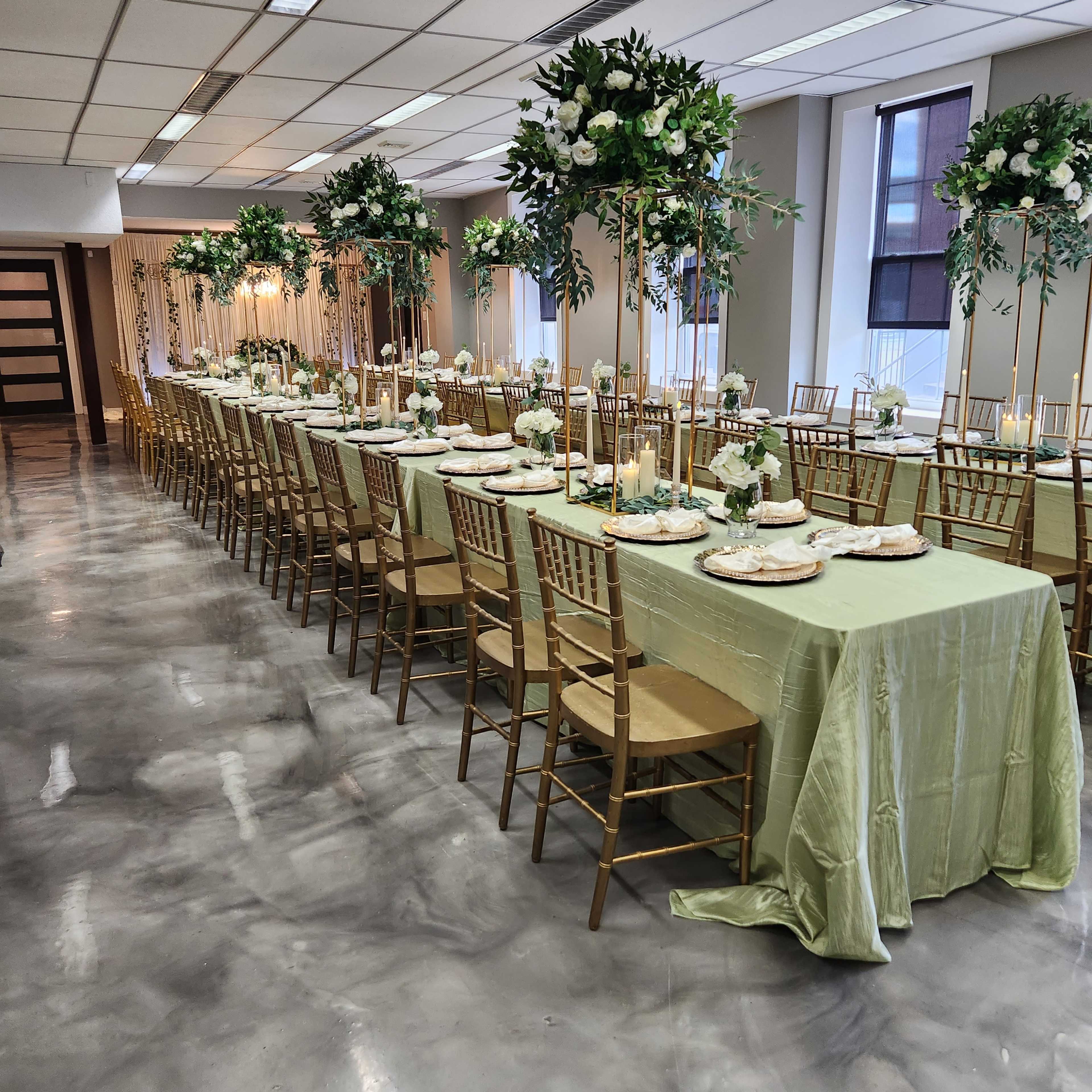 A long banquet table set for an event with gold chairs, floral centerpieces, and green tablecloths.