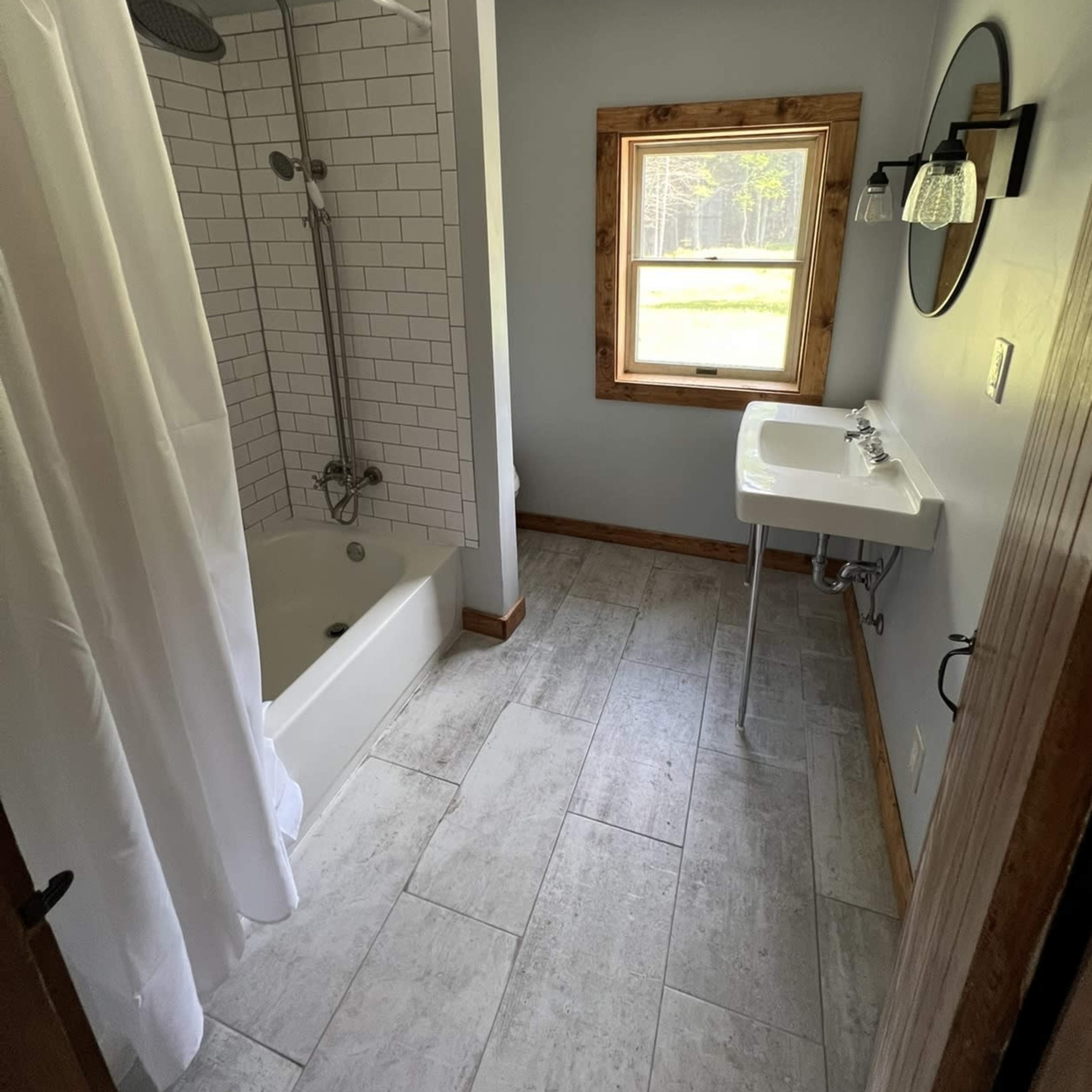 A small bathroom featuring a shower with a white curtain, a sink mounted on a pedestal, and a window allowing natural light.