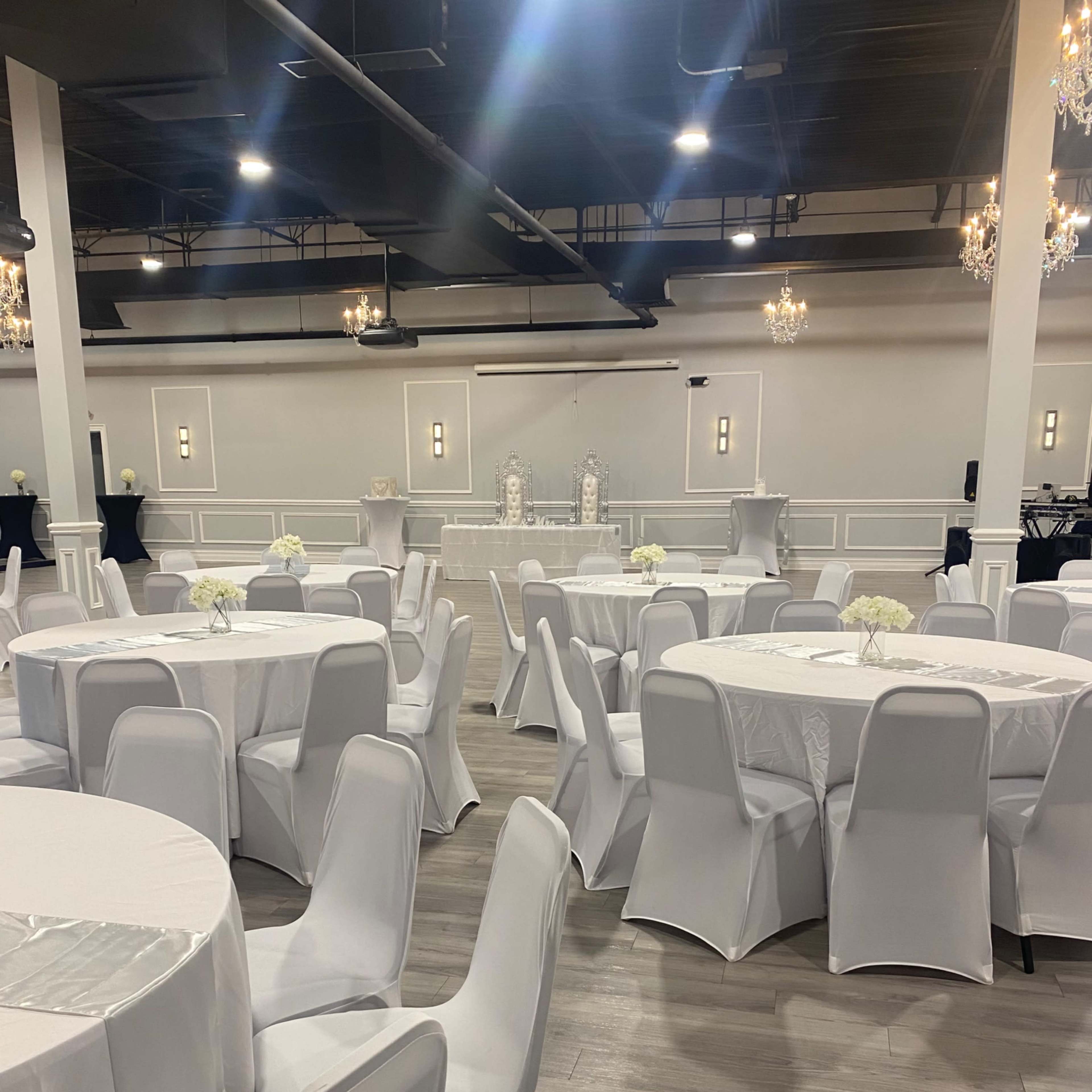 A large event space with round tables covered in white tablecloths and white chairs, arranged in a setup for an event, with chandeliers hanging above.