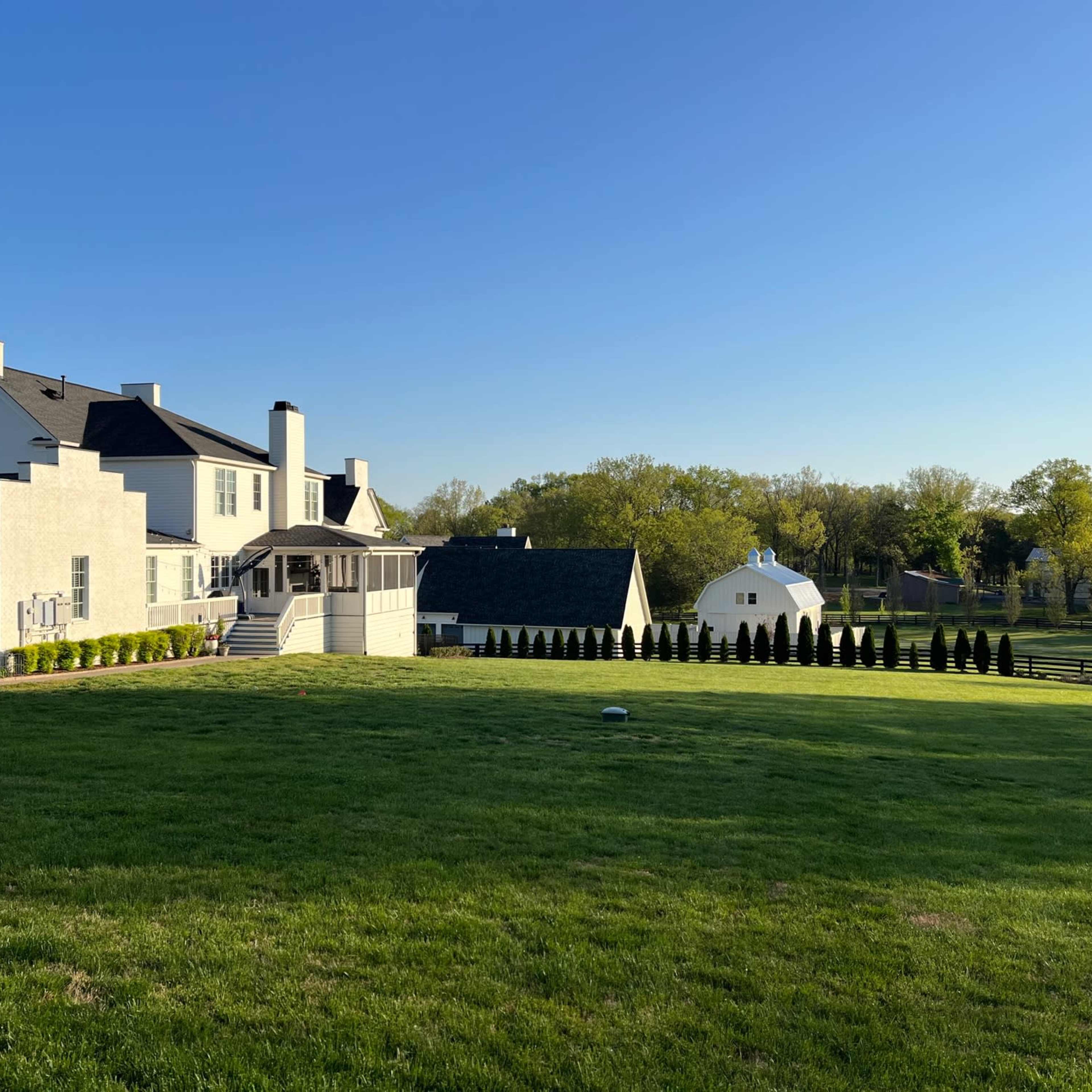 A spacious lawn surrounds a modern white house and a nearby barn under a clear blue sky.