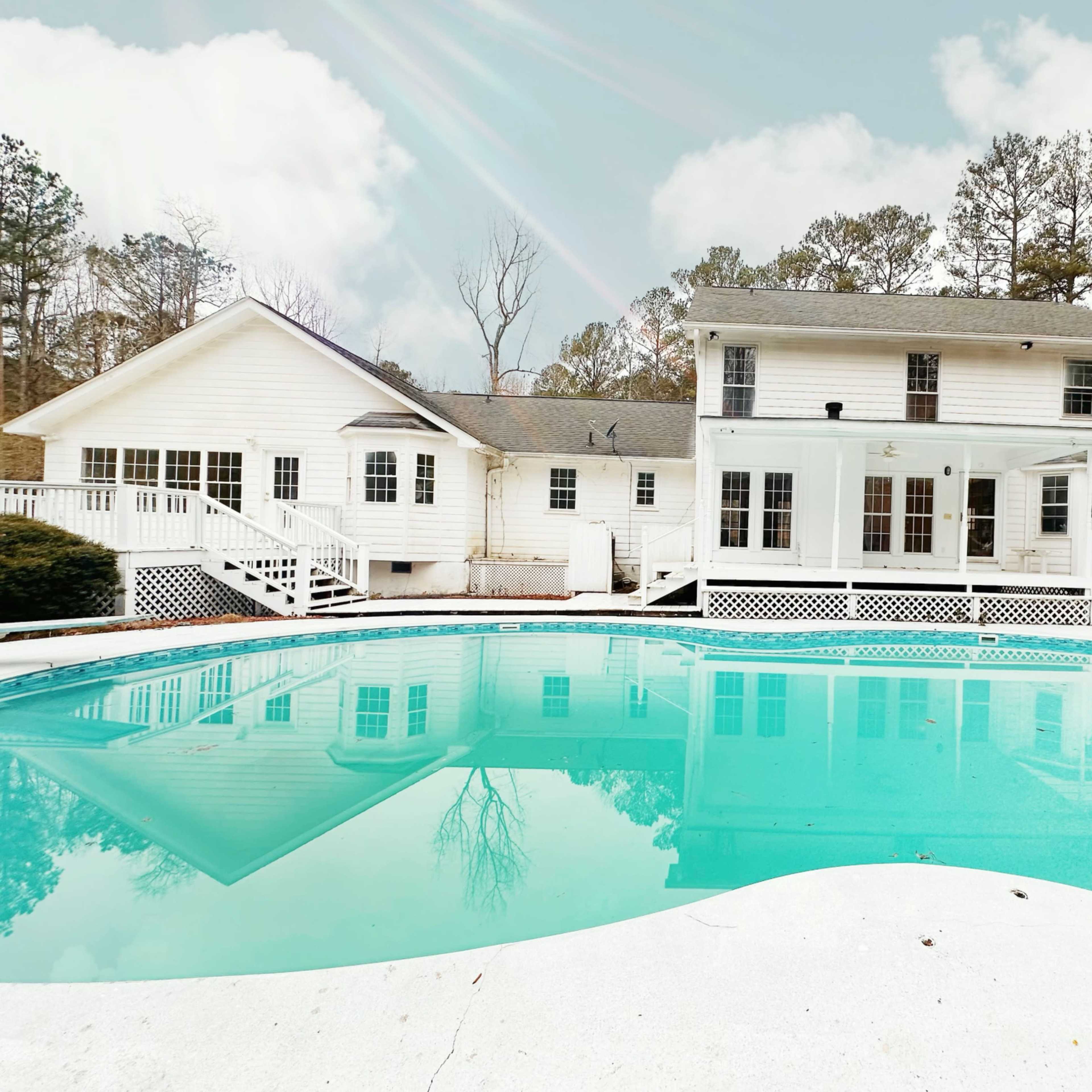 A large, residential home with multiple sections surrounds a turquoise swimming pool, set among tall trees.