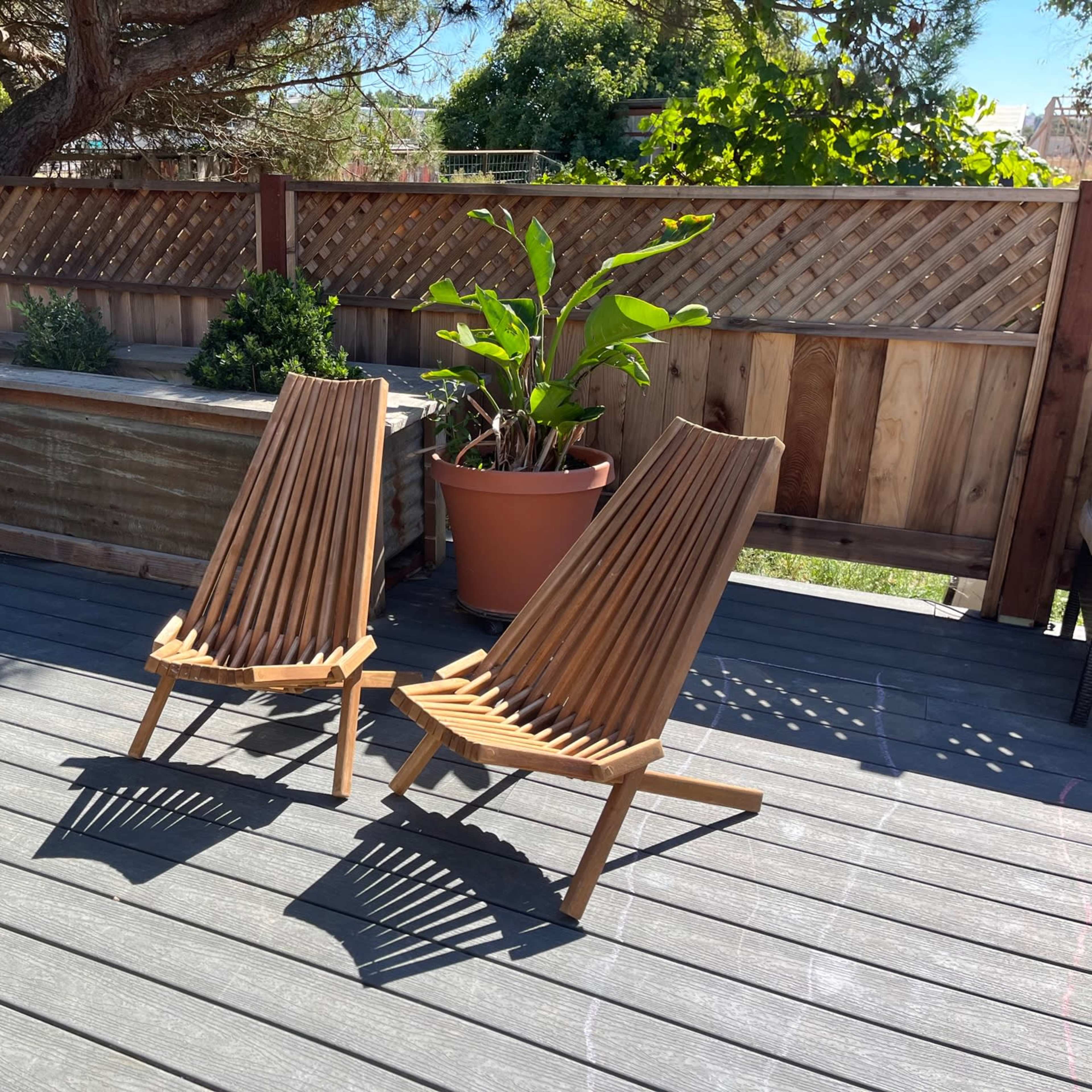 Two wooden slatted chairs are positioned on a deck beside a planter with green plants and a wooden fence in the background.