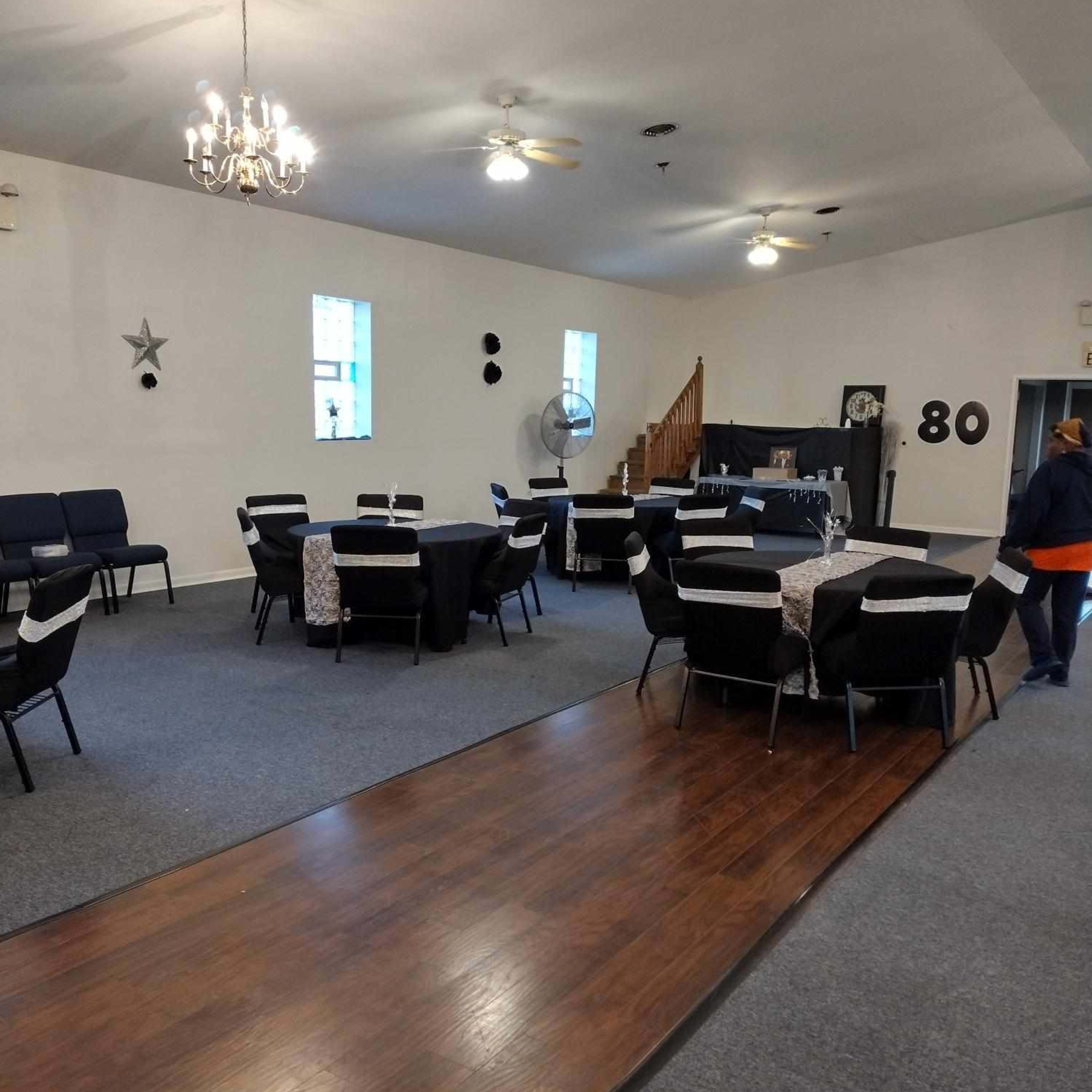 A spacious event room arranged with tables and chairs, featuring a wooden floor section and decorative elements like a chandelier and wall decor.