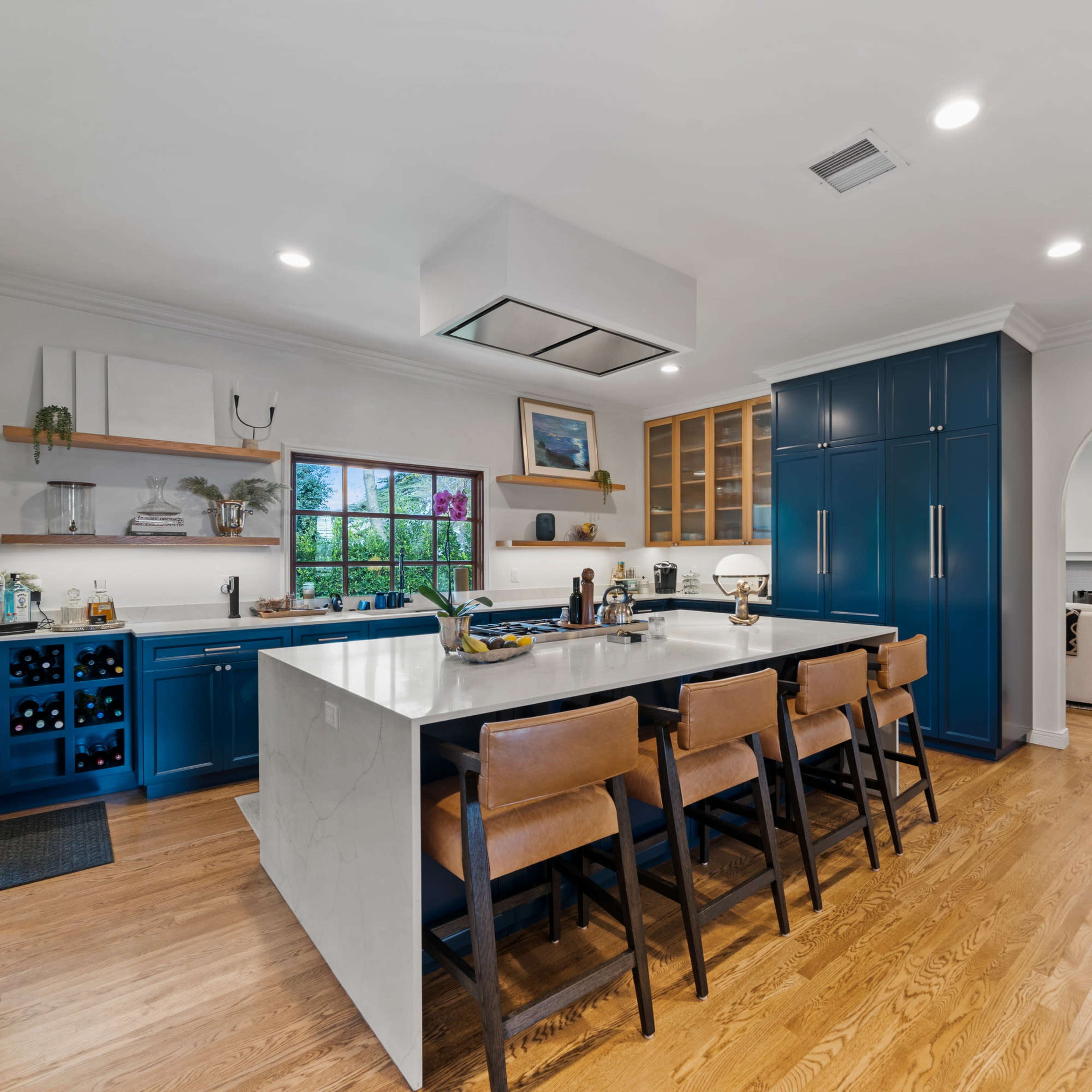 A modern kitchen features blue cabinetry, a large island with seating, and a view of an adjoining living area.