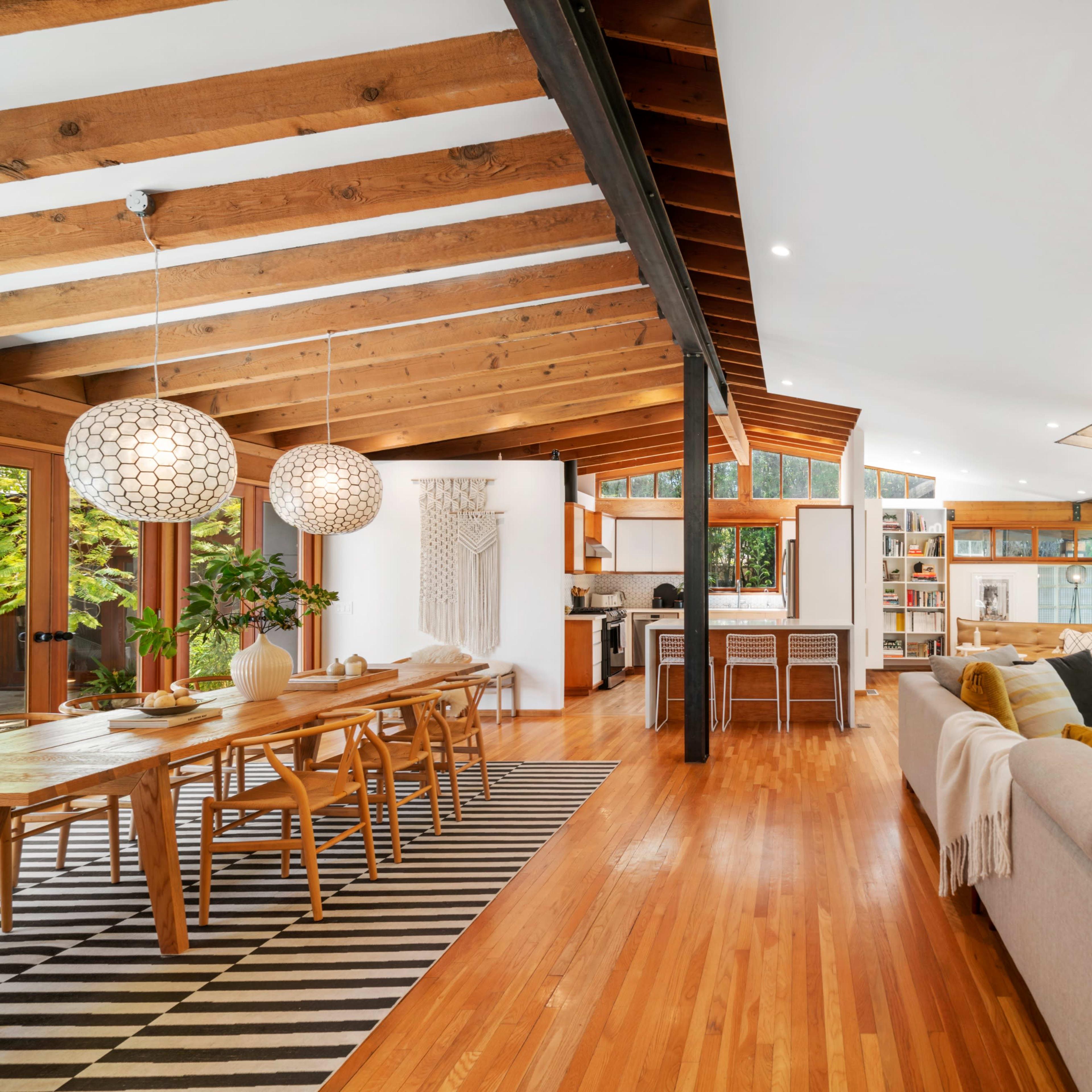 An open-concept living area with wooden beams, a dining table, and a comfortable sofa, surrounded by large windows allowing natural light.