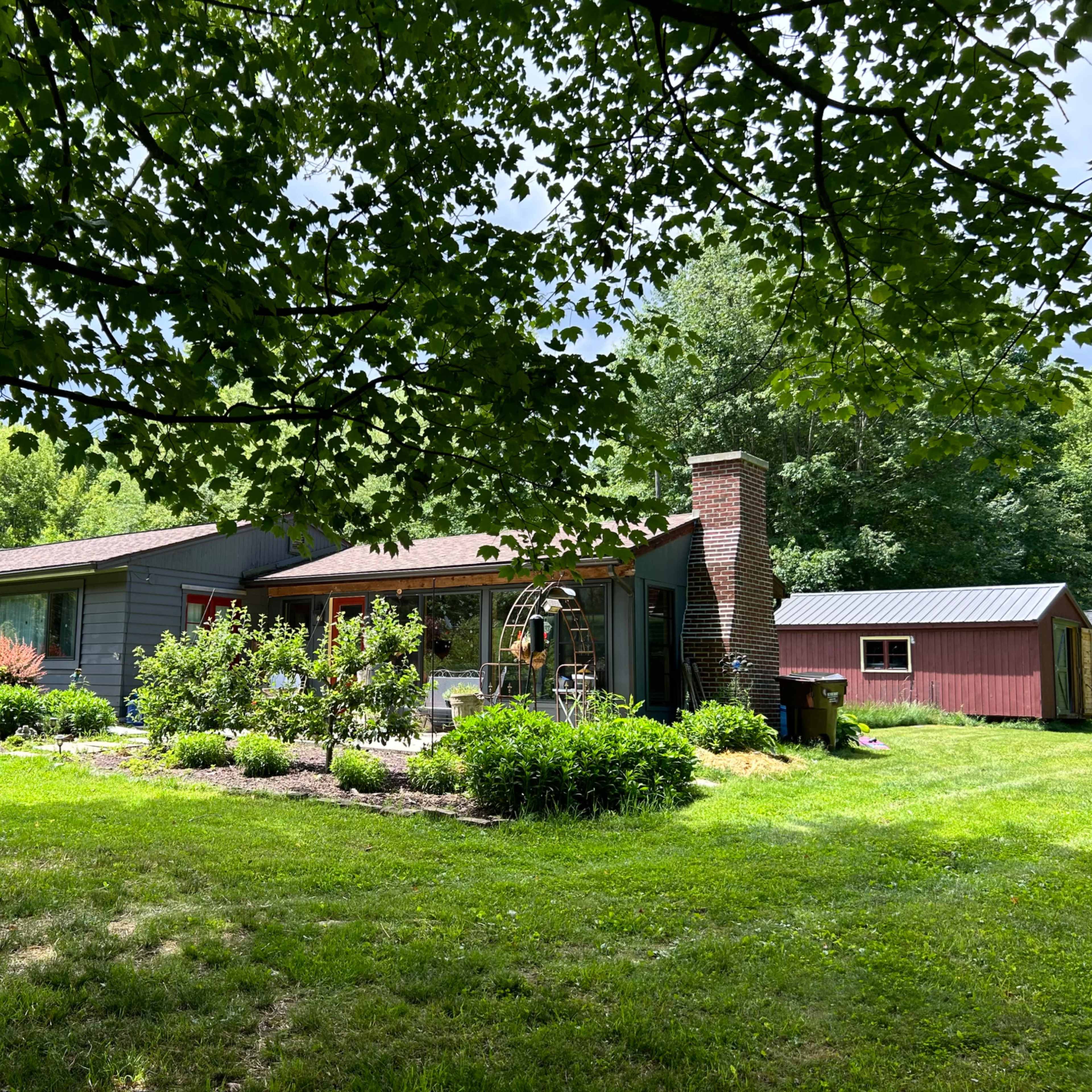 A single-story gray house with a red door and a brick chimney is surrounded by lush greenery and a small red shed in the background.