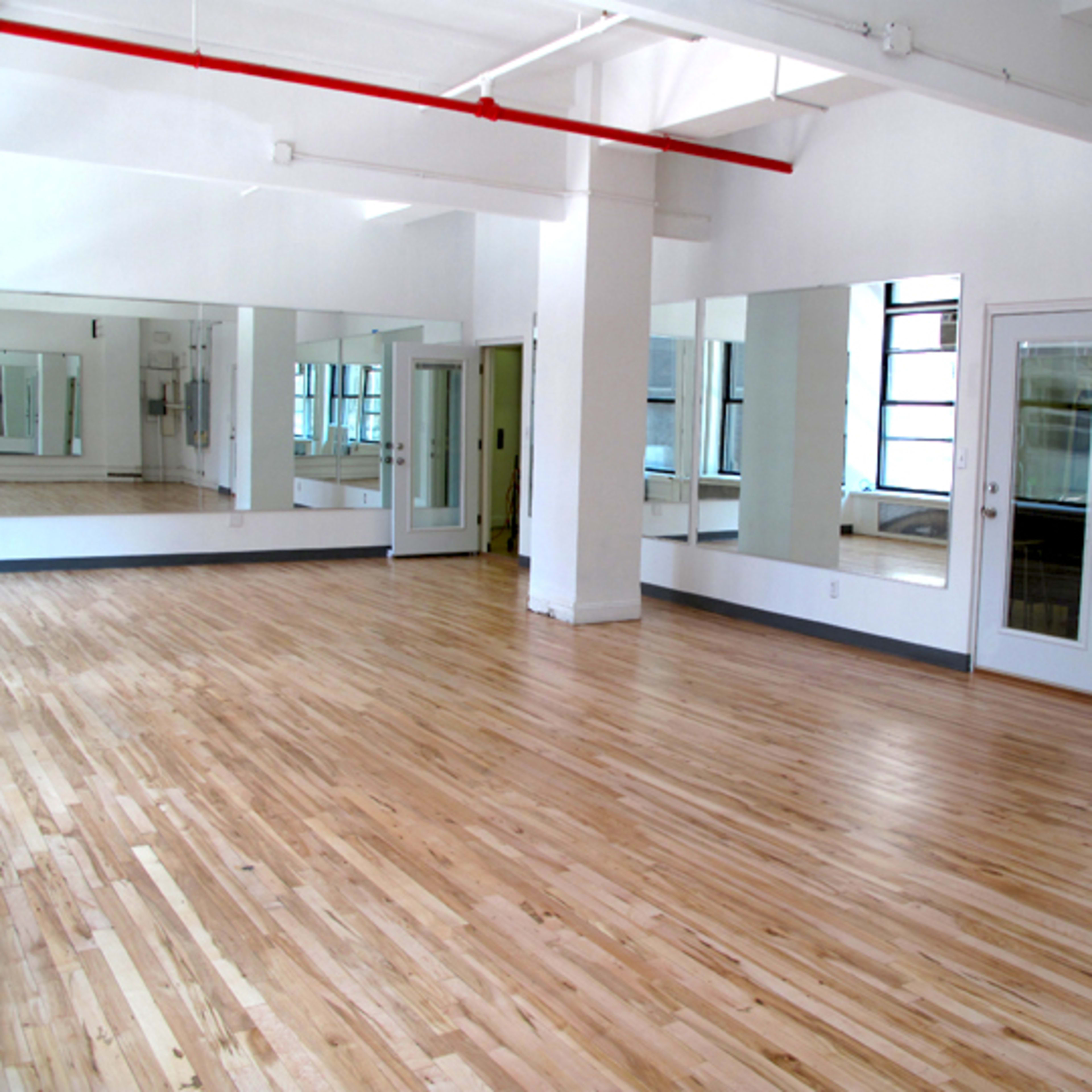 An empty studio space with wooden flooring, large mirrors on one wall, and double doors leading to another room.