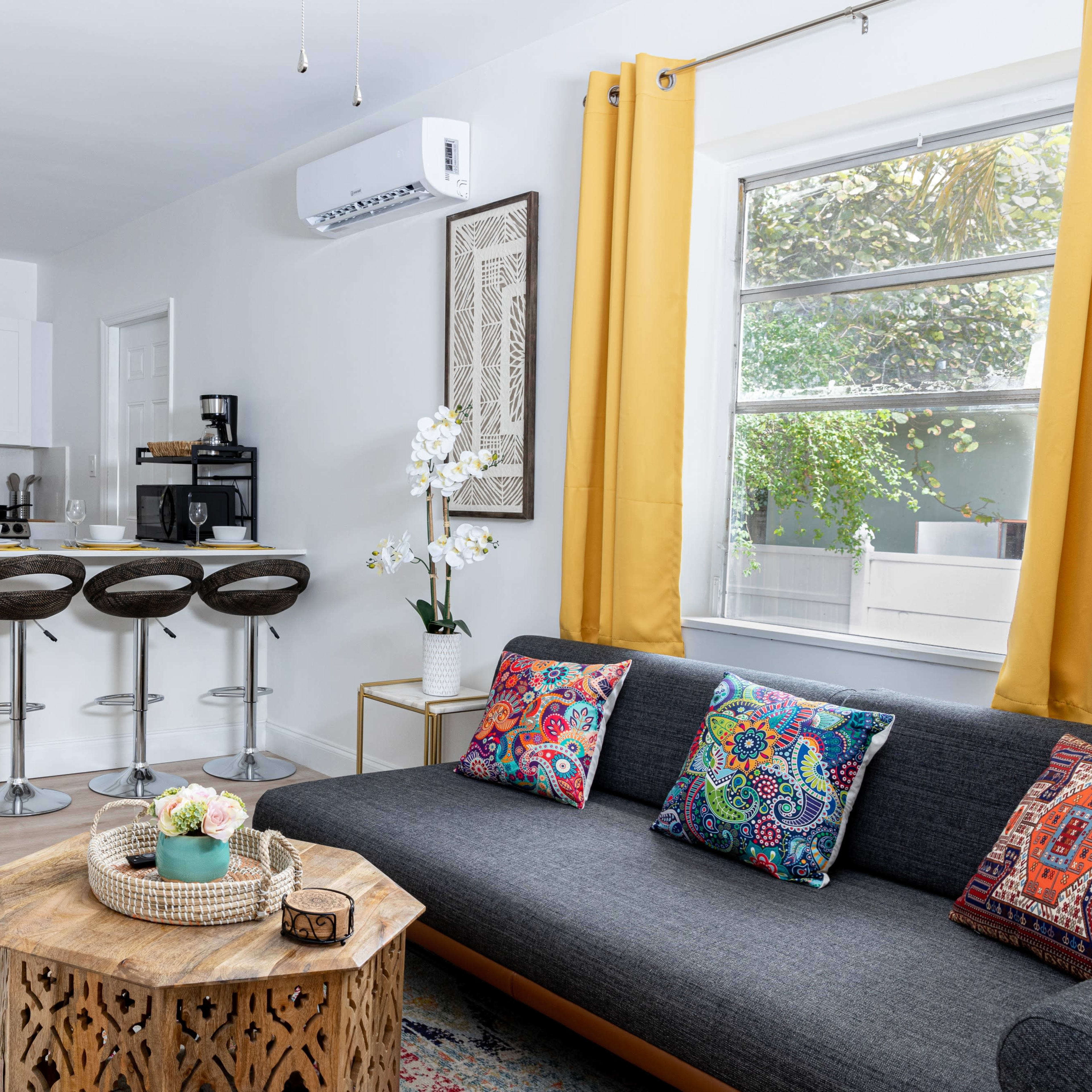 A modern living space features a gray sofa with colorful cushions, a wooden coffee table, and a kitchen area with bar stools and stainless steel appliances.