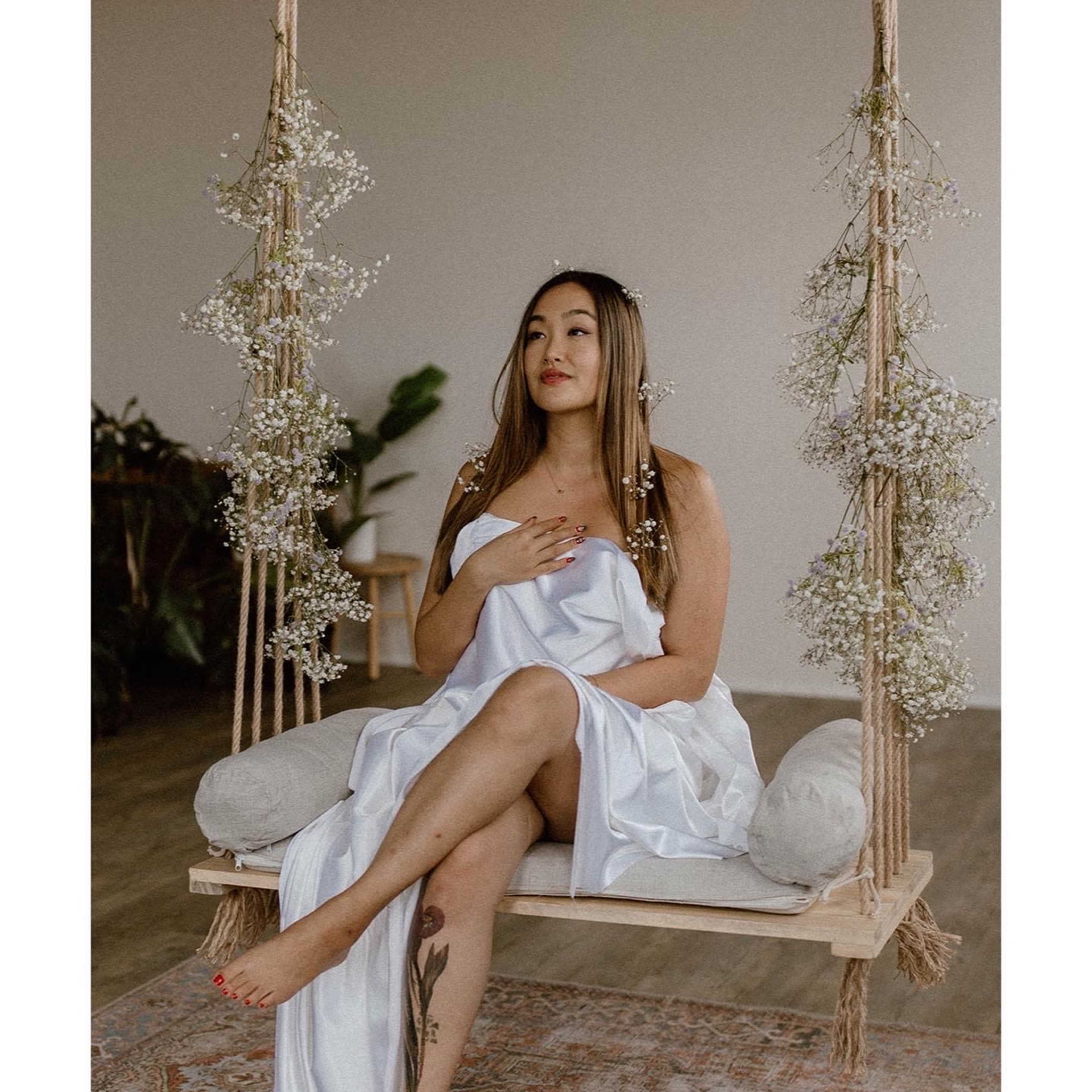A woman sits on a wooden swing adorned with flowers, wearing a white dress, in a softly lit room.