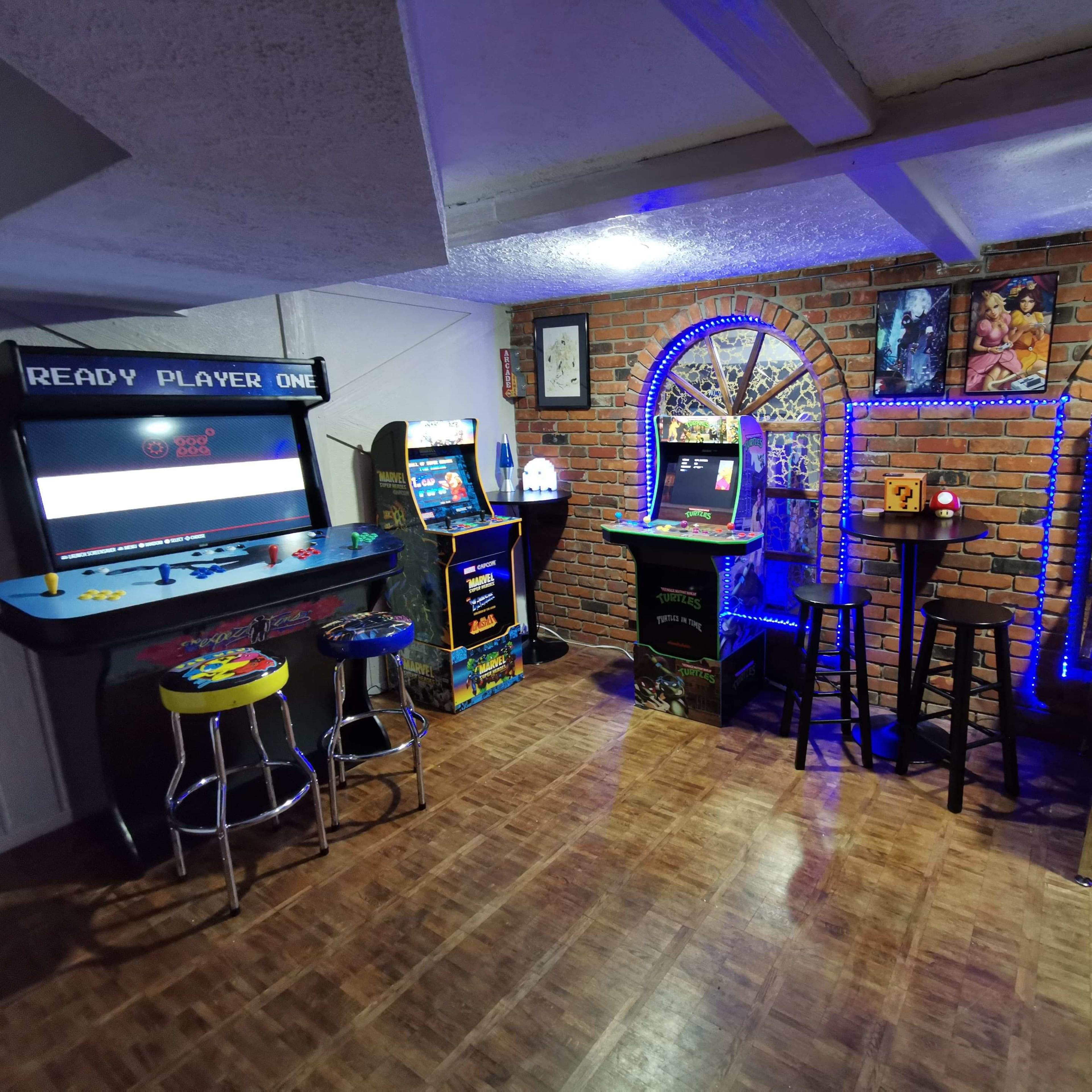 A gaming room with various arcade machines, a pinball machine, and colorful lighting against a brick wall.